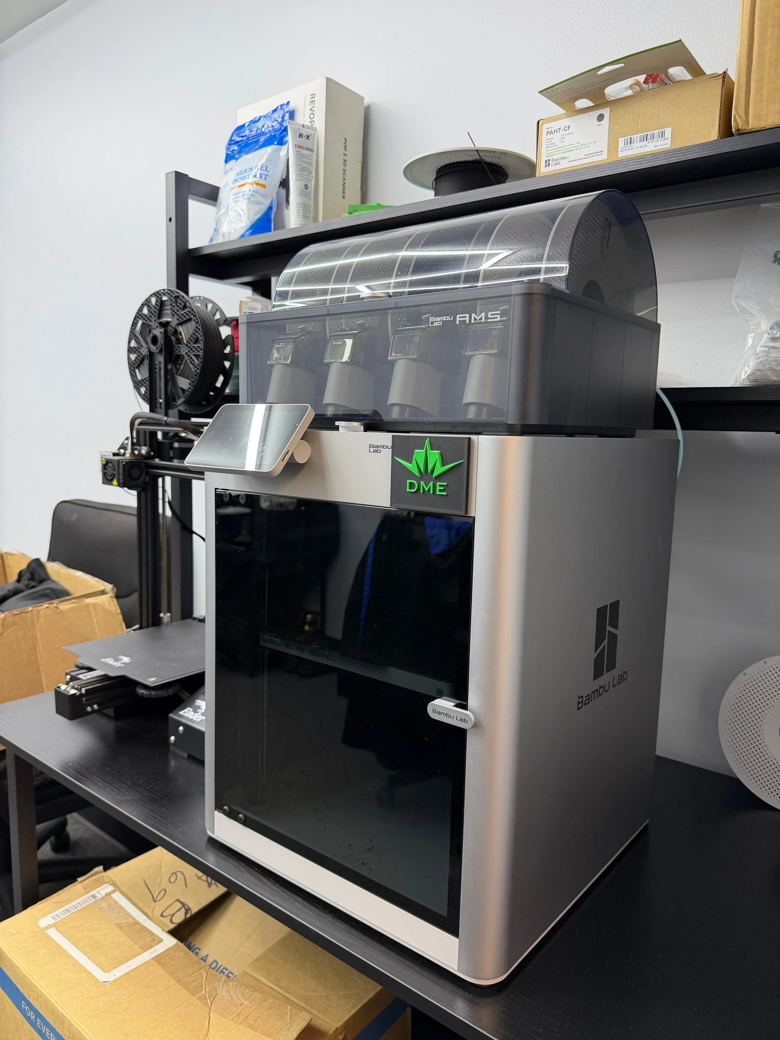 A Bambu Lab 3D printer with a touchscreen interface, situated on a black worktable with boxes and shelves in the background.