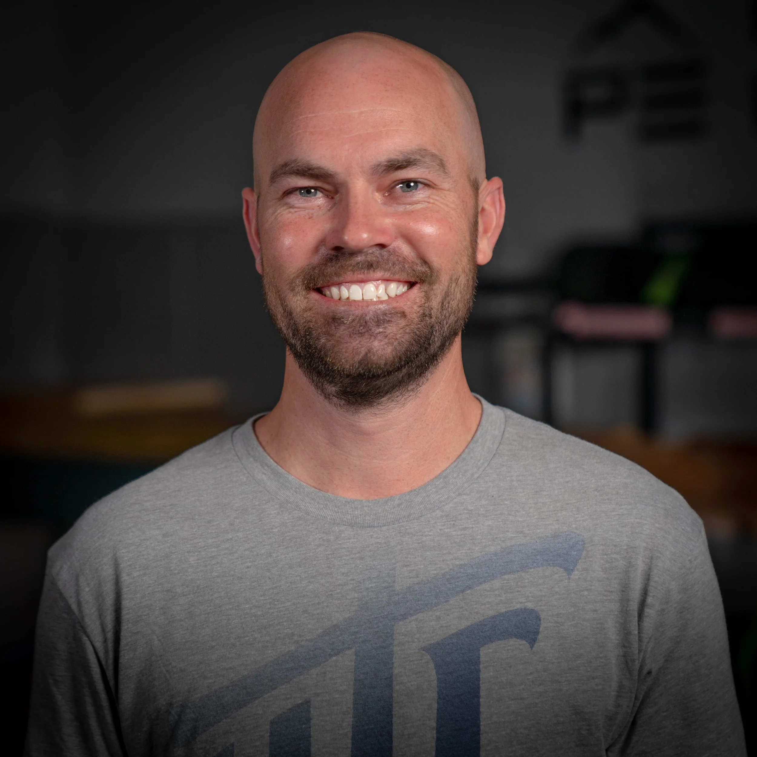 Close-up of a smiling man with a shaved head and light-colored eyes, wearing a gray t-shirt.