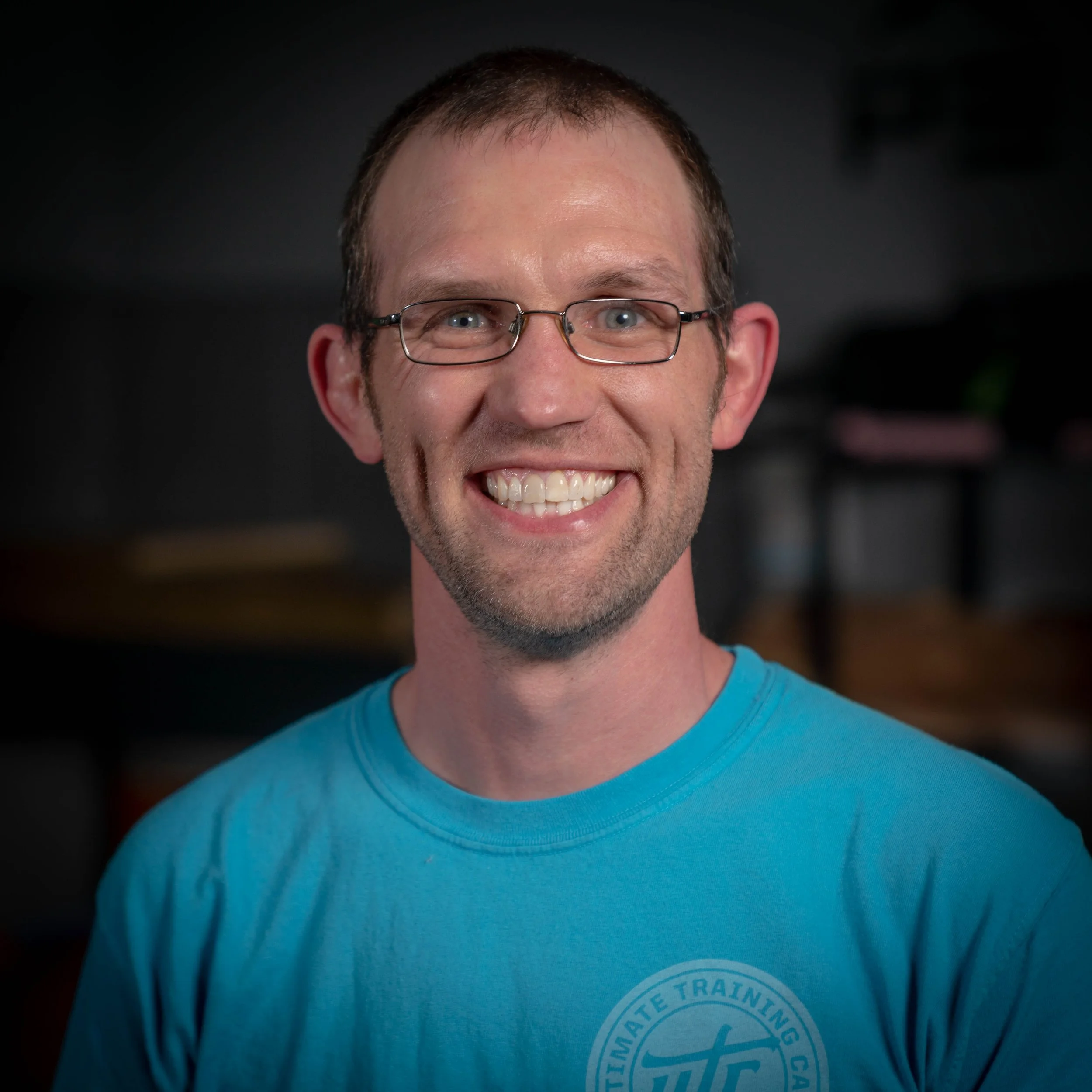 A smiling man with glasses, short brown hair, and a light complexion, wearing a blue t-shirt with a circular logo that reads 'Ultimate Training'. He is indoors with a dark background.