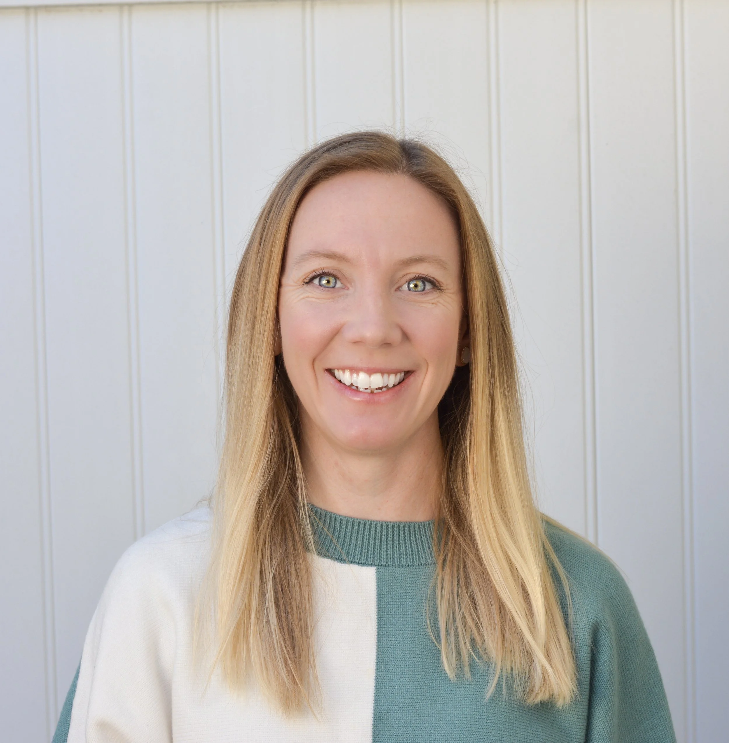 A smiling woman with long blonde hair and blue eyes standing against a light-colored paneled wall.