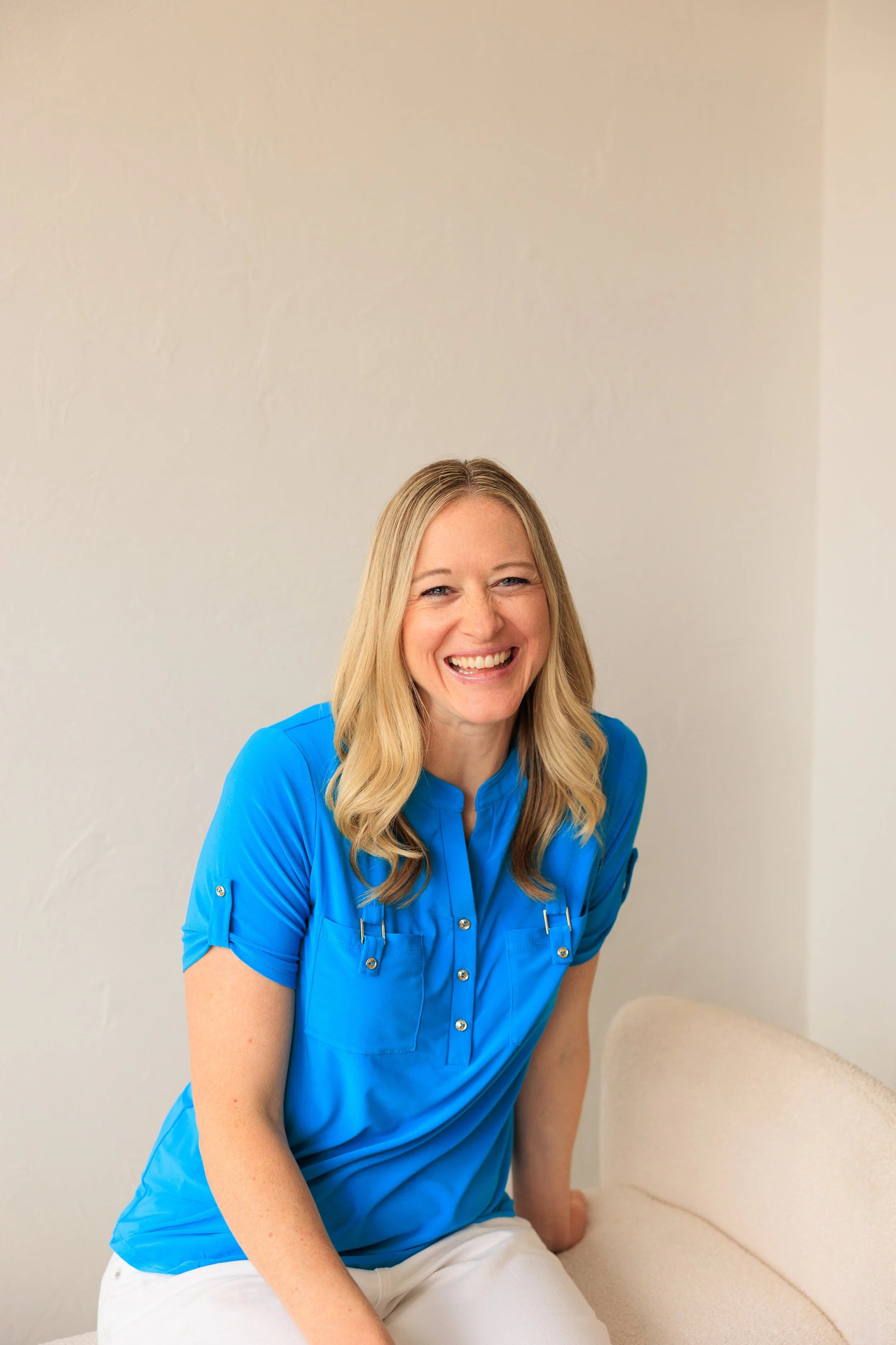 Emily Ricks, Life coach and founder of Think New Thoughts Academy  with blonde hair smiling, wearing a blue medical scrubs top, sitting on a beige couch against a plain light-colored wall.