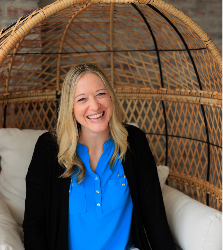 Emily Ricks, Certified Life Coach with long blonde hair smiling, sitting inside a wicker cocoon chair with a brick wall background, wearing a blue blouse and black jacket.