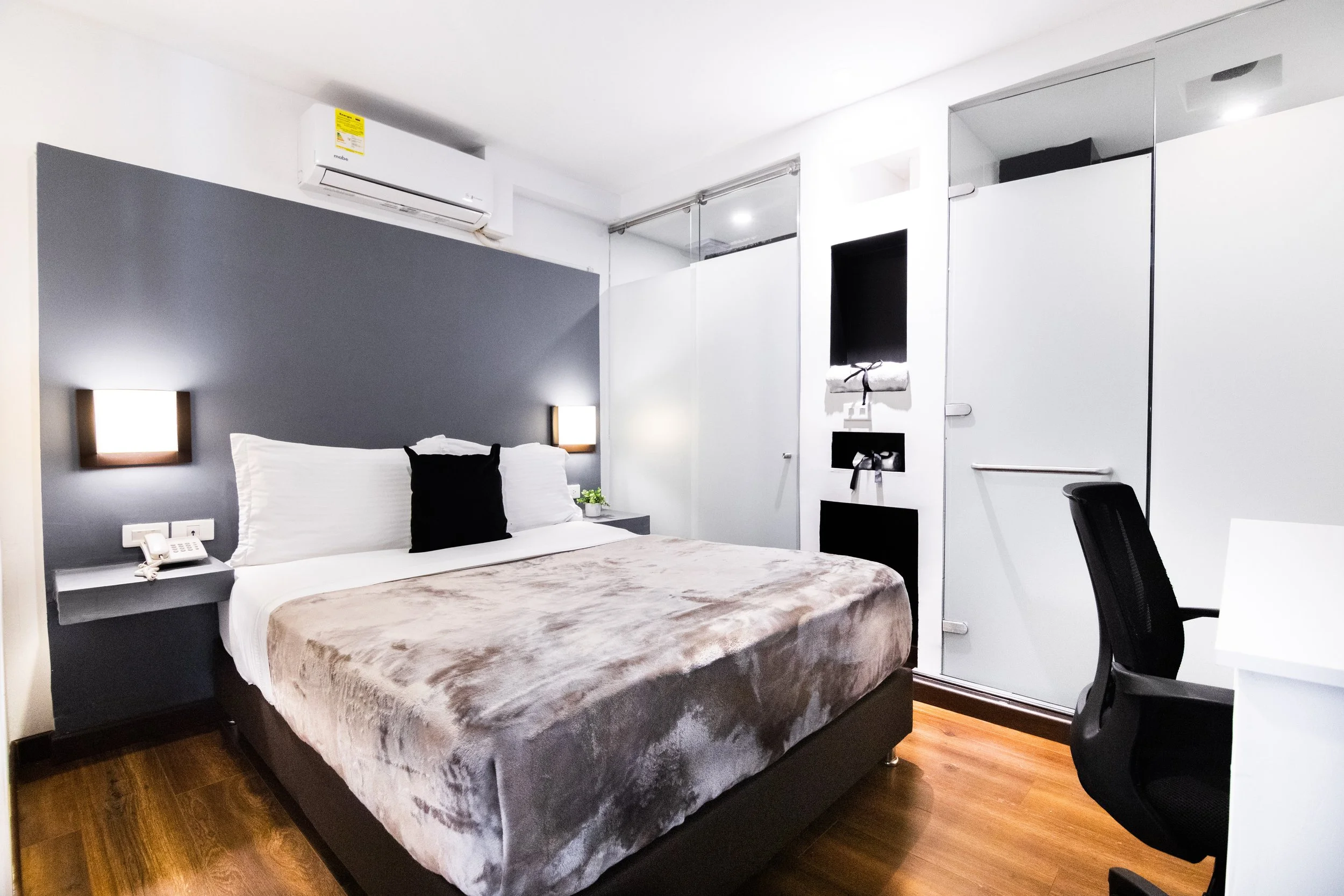 Habitación moderna con cama grande, paredes blancas y grises, lámparas de lectura, aire acondicionado, escritorio y silla negra.