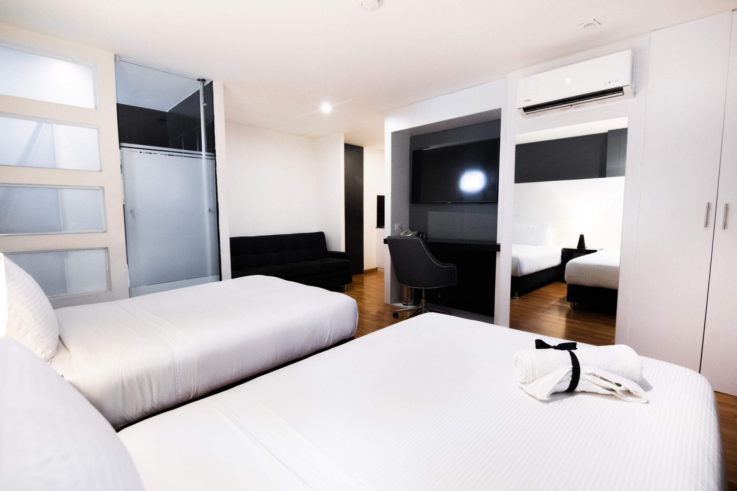 Habitación moderna con dos camas blancas, un sofá negro, televisión grande y área de trabajo, piso de madera y paredes blancas.