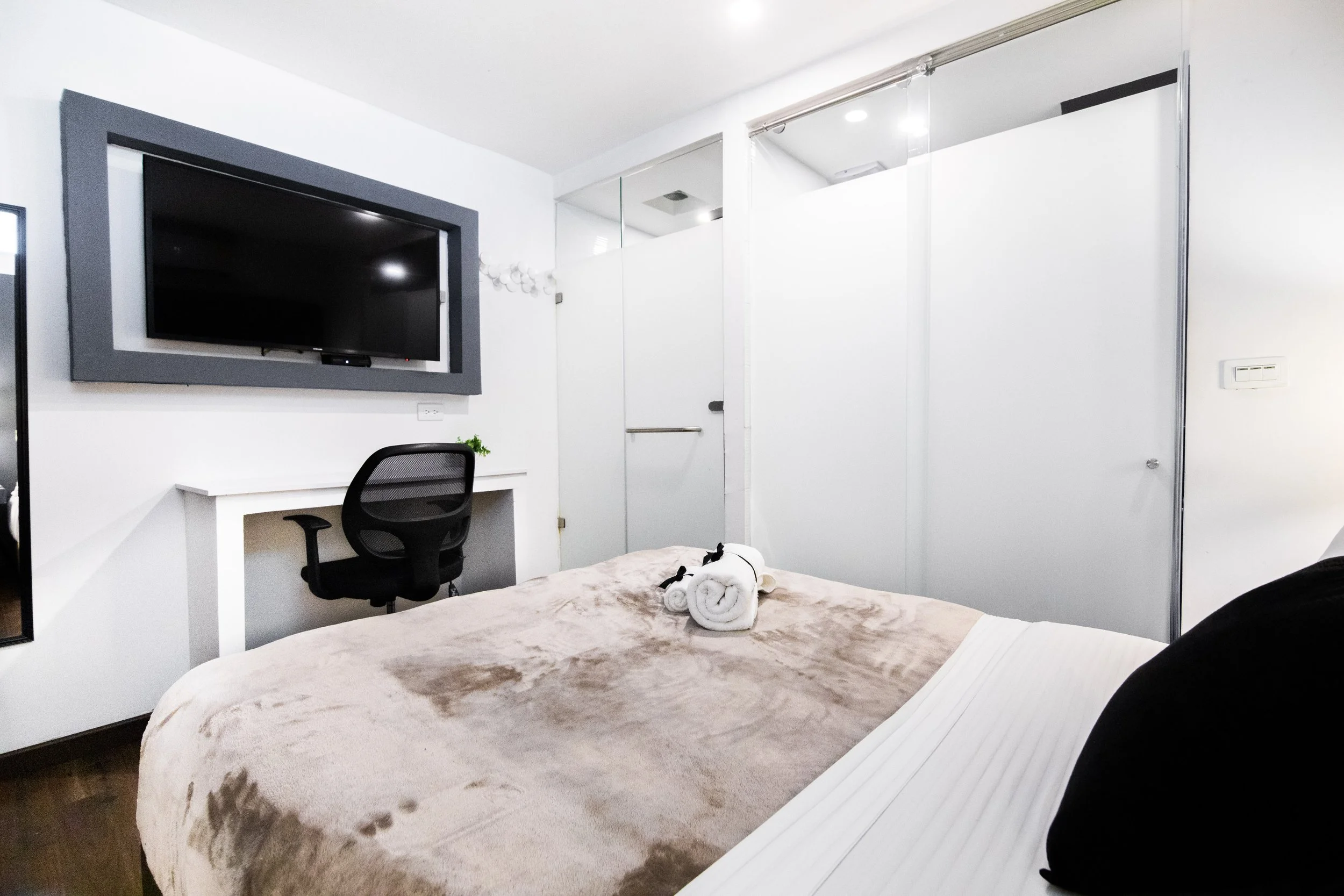 Habitación moderna con cama, toallas enrolladas, televisor en la pared, escritorio con silla y puertas corredizas de vidrio.
