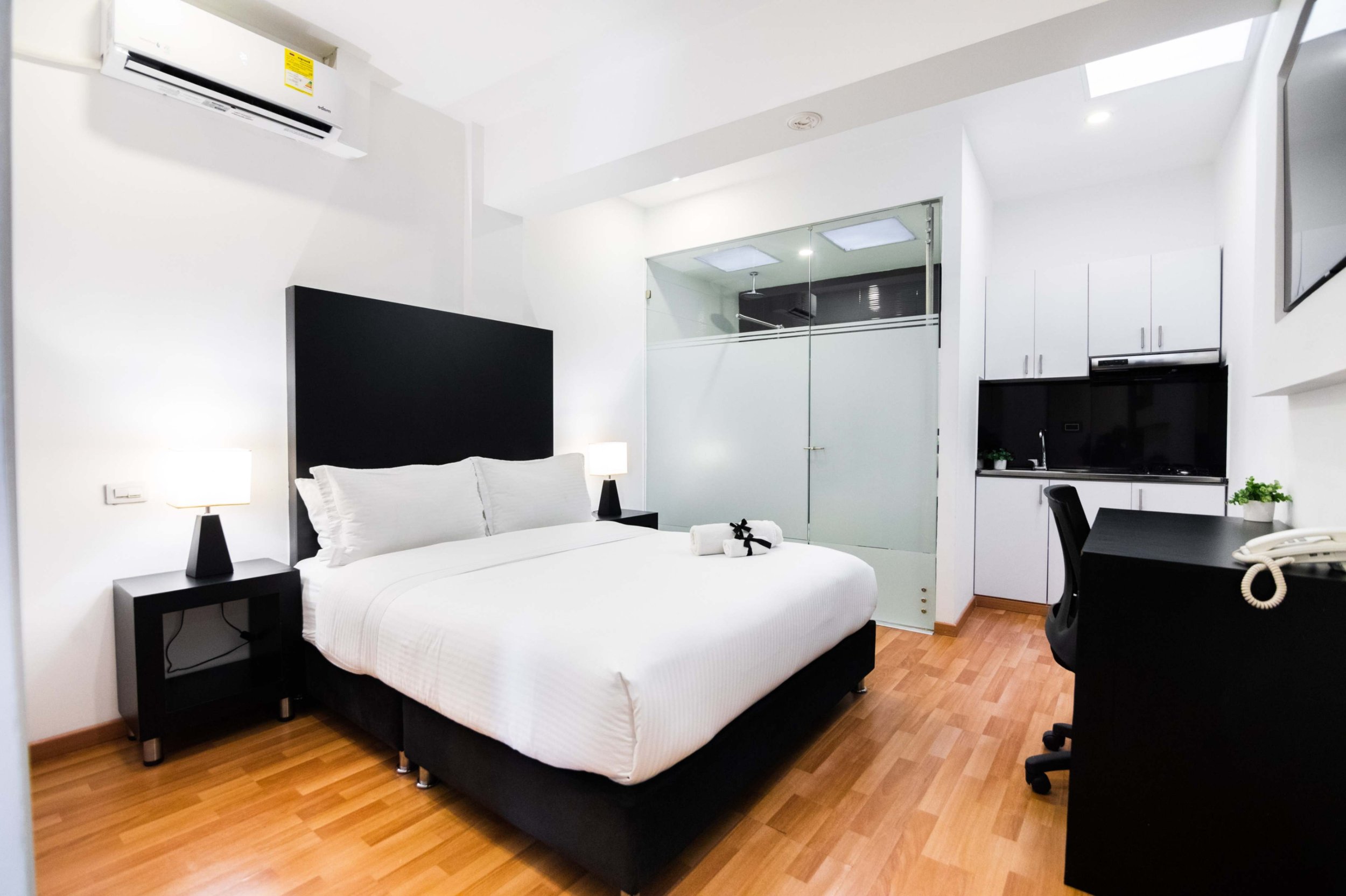 Habitación de hotel con cama grande, dos lámparas de noche, aire acondicionado en la pared, escritorio con silla y micrófono, cocina pequeña y puerta de vidrio con cortinas.