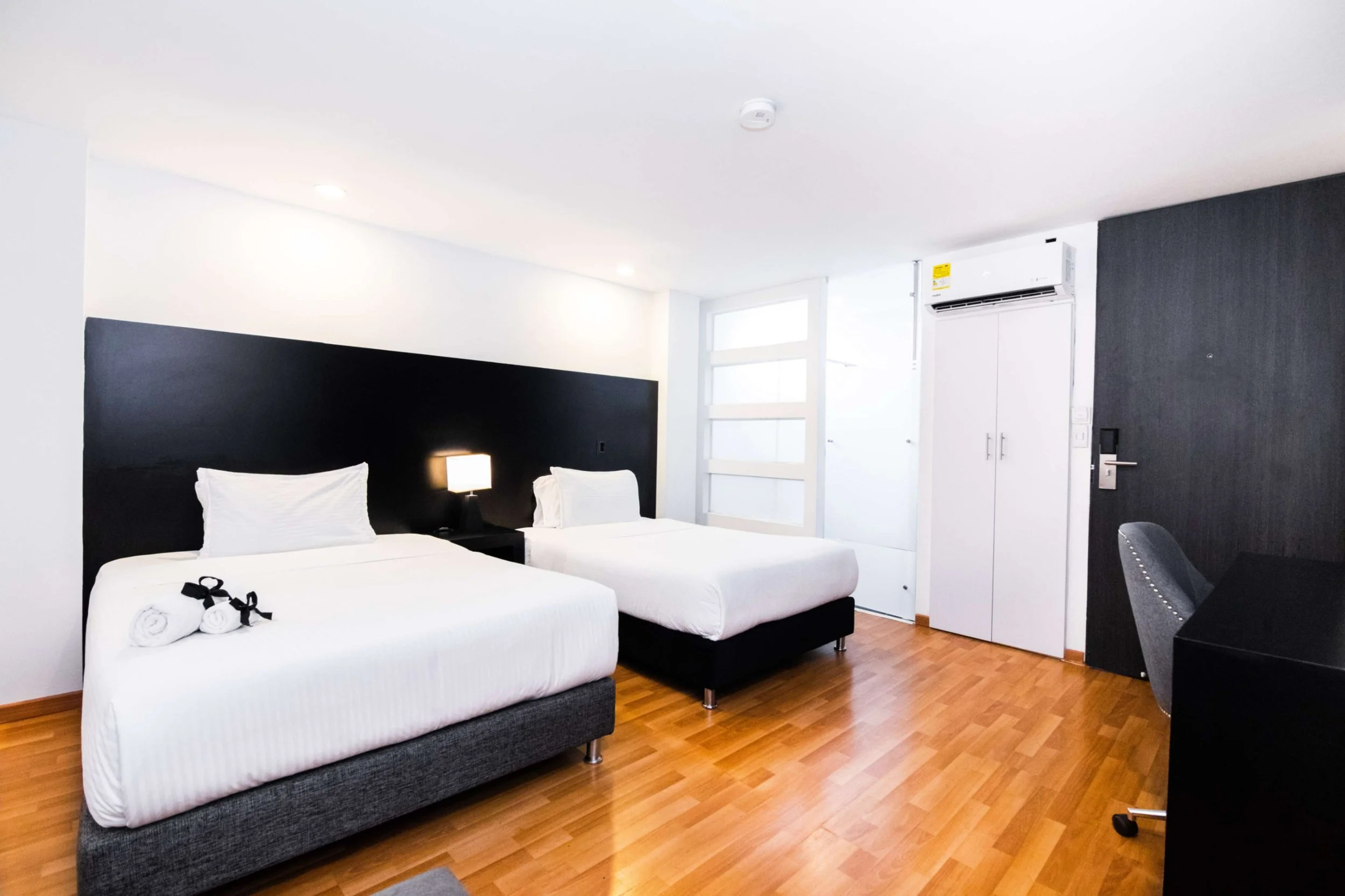 Habitación moderna con dos camas individuales con ropa de cama blanca, una lámpara en la cabecera, piso de madera, paredes blancas y una pared en tono oscuro, aire acondicionado, puerta de vidrio con paneles, armario blanco y mesa de trabajo con sill