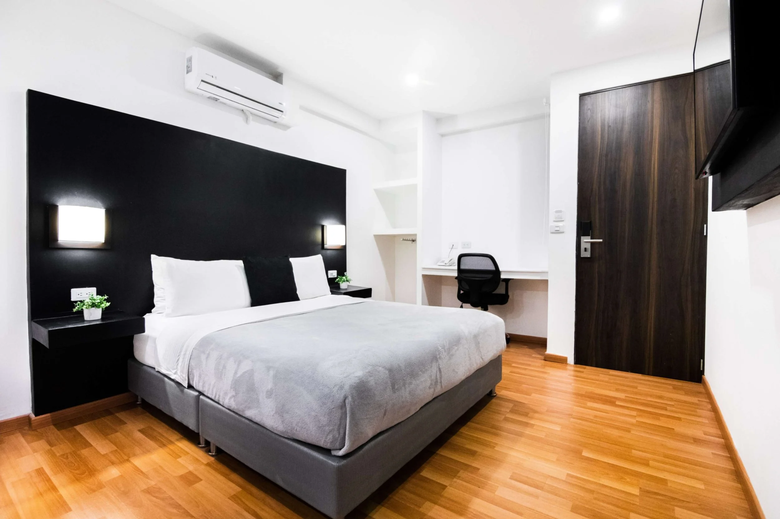 Habitación moderna con cama grande, paredes blancas, suelo de madera, televisor en la pared, escritorio con silla y aire acondicionado.