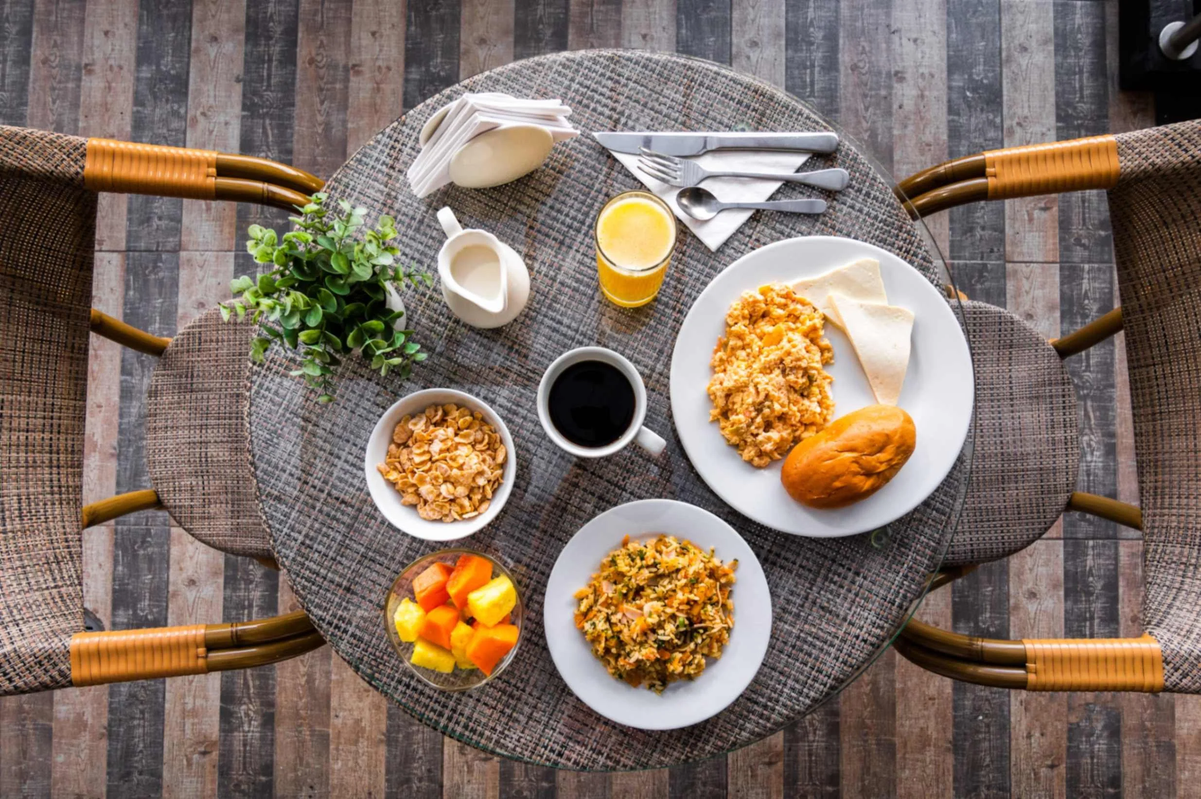 Desayuno en una mesa redonda con huevo revuelto, cereal, café, jugo de naranja, pan, tortilla, y frutas en un plato. Hay cubiertos y una jarra de leche.