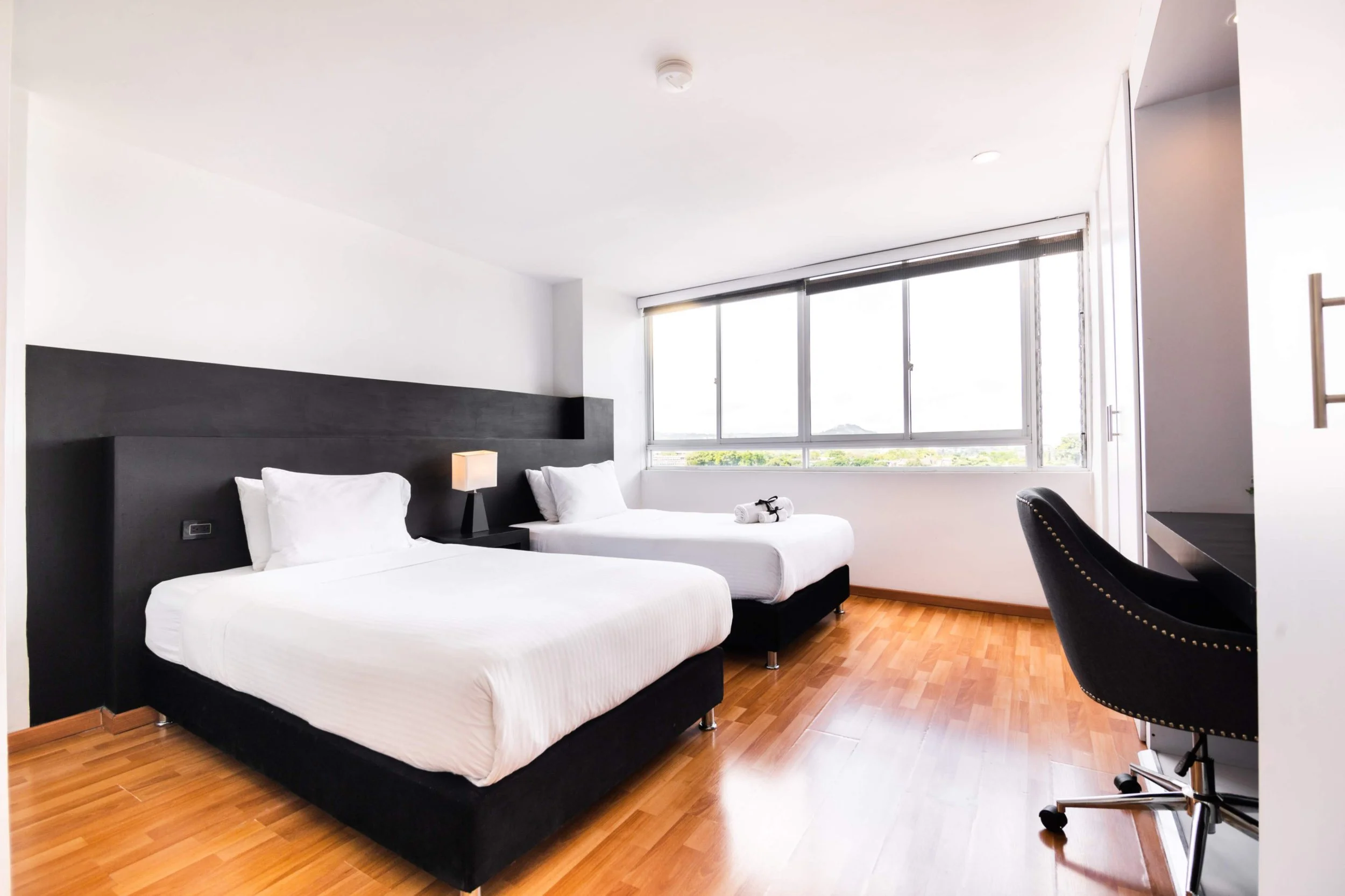 Habitación de hotel con dos camas con sábanas blancas, una lámpara entre ellas, vista a través de una ventana grande con persiana, suelo de madera y escritorio con silla negra.