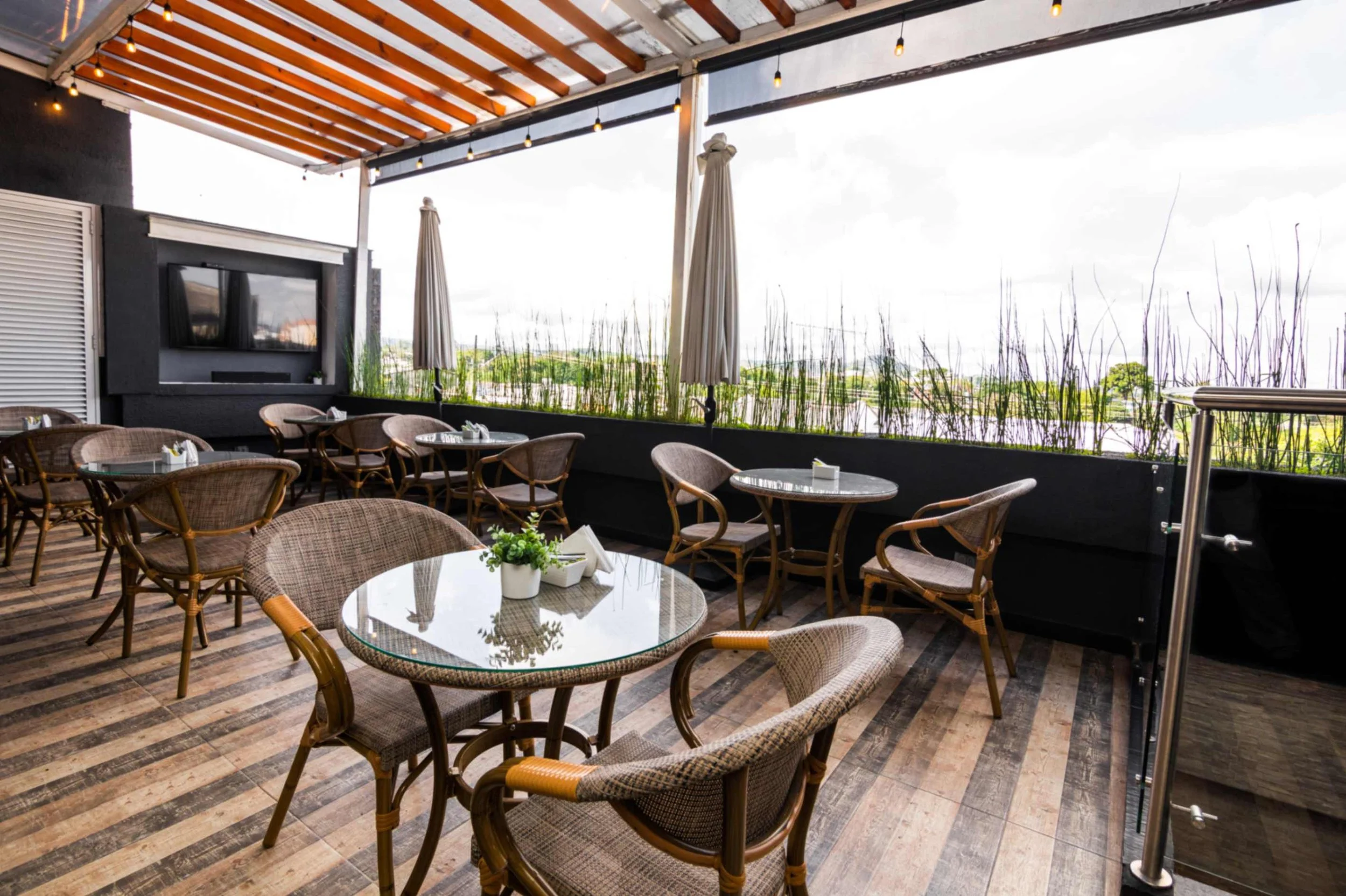 Terraza de restaurante al aire libre con mesas de vidrio y sillas de mimbre, decorada con plantas y protegida por un techo de tablones de madera.