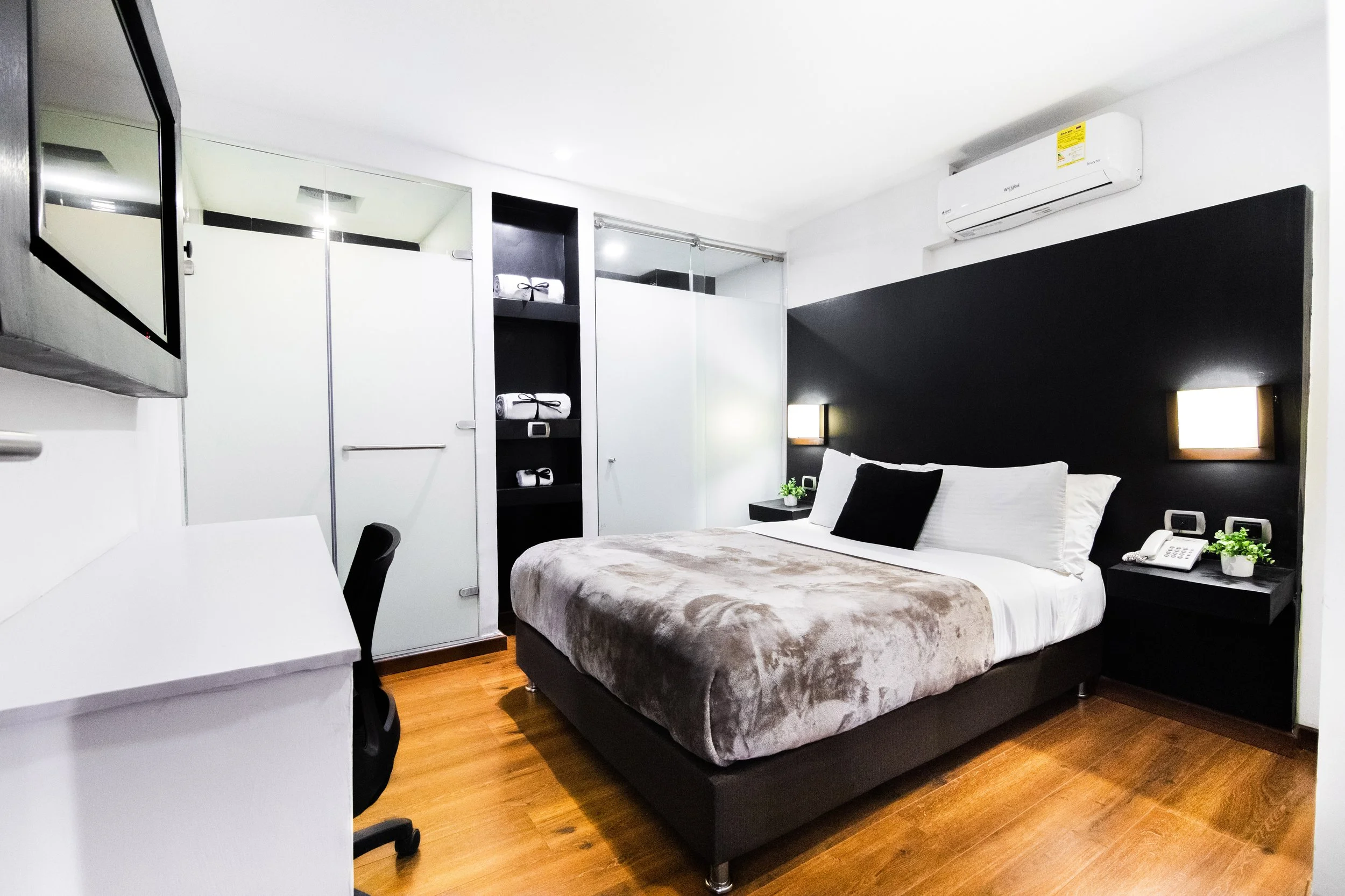 Habitación de hotel con cama doble, paredes blancas, ventanales con cortinas blancas, escritorio blanco, silla negra, armario oscuro con toallas, murales en negro y blanco, televisor en la pared, aire acondicionado y lámparas de noche