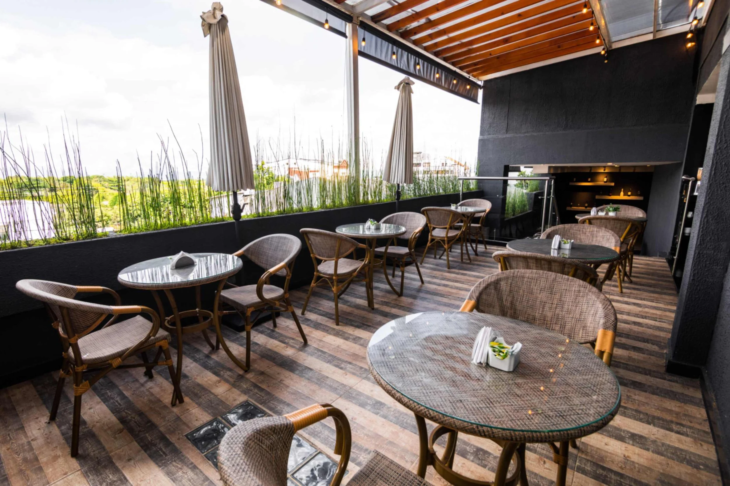 Terraza de restaurante con mesas redondas de vidrio, sillas de mimbre, sombrillas cerradas y piso de madera, decorada con plantas en el borde.
