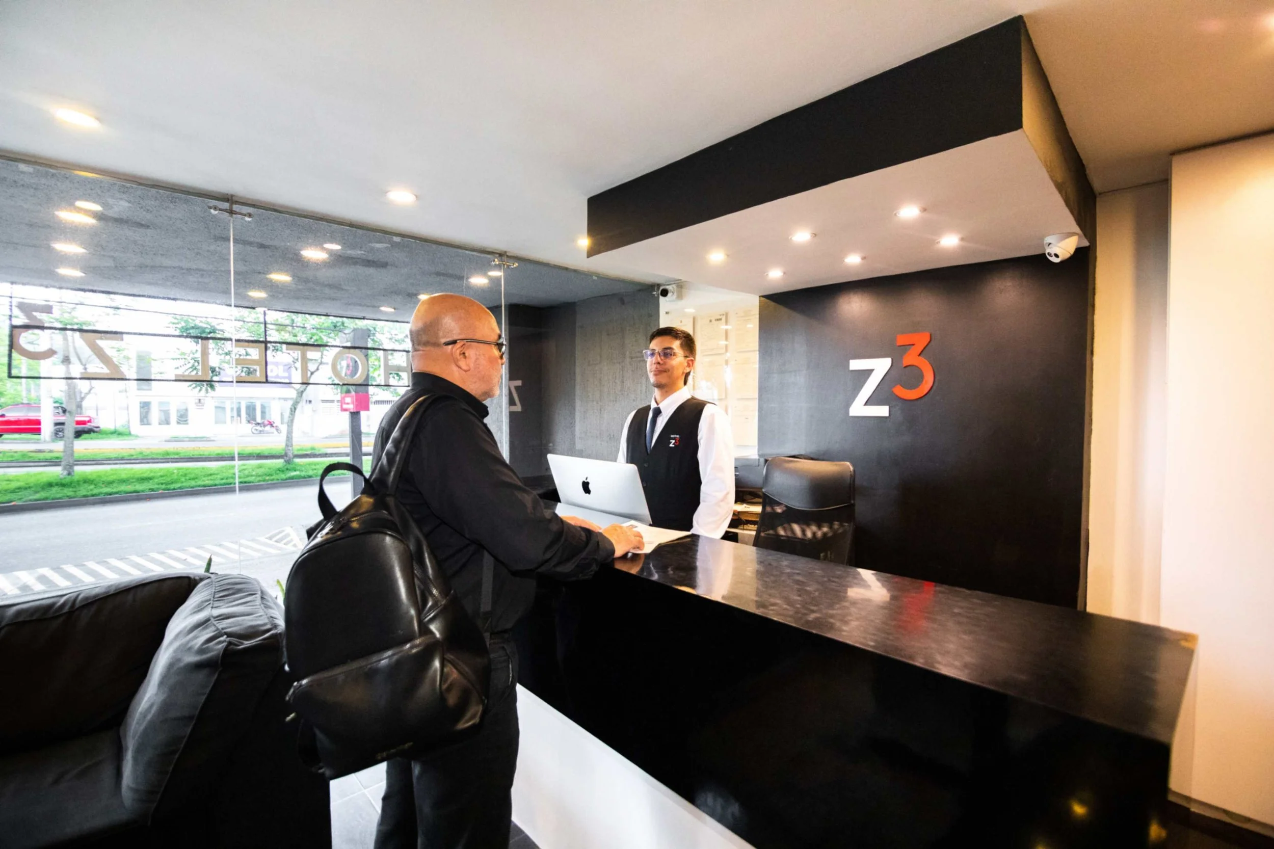 Recepción de un hotel con un hombre atendiendo a un cliente, mostrando el logo "z3" en la pared.
