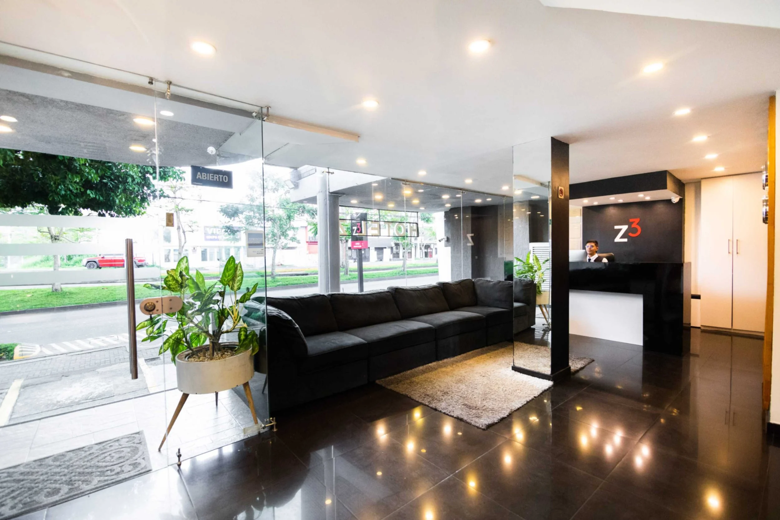 Recepción de una oficina moderna con un sofá negro, plantas decorativas y un mostrador con el logotipo "Z3". La entrada es de vidrio y un letrero indica "Abierto".
