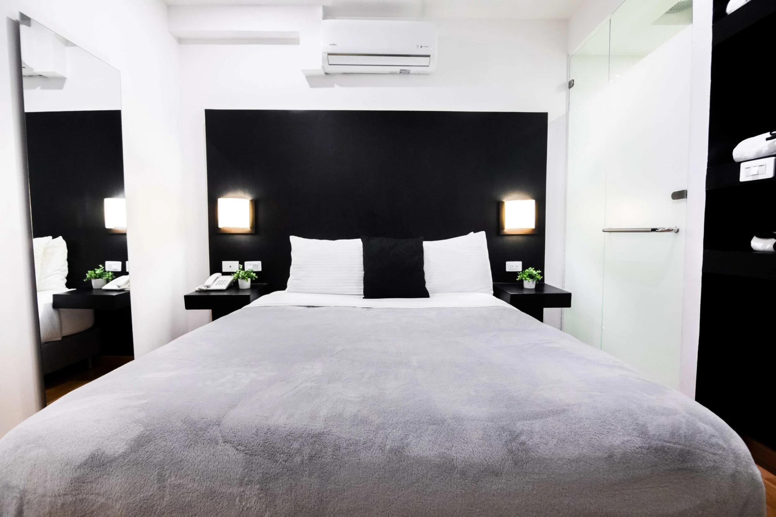 Cama king size con cabecera negra, almohadas blancas y una almohada negra en el centro, lámparas de pared con luz blanca, mesas de noche negras con plantas verdes y teléfonos, paredes blancas y un aire acondicionado en la pared.