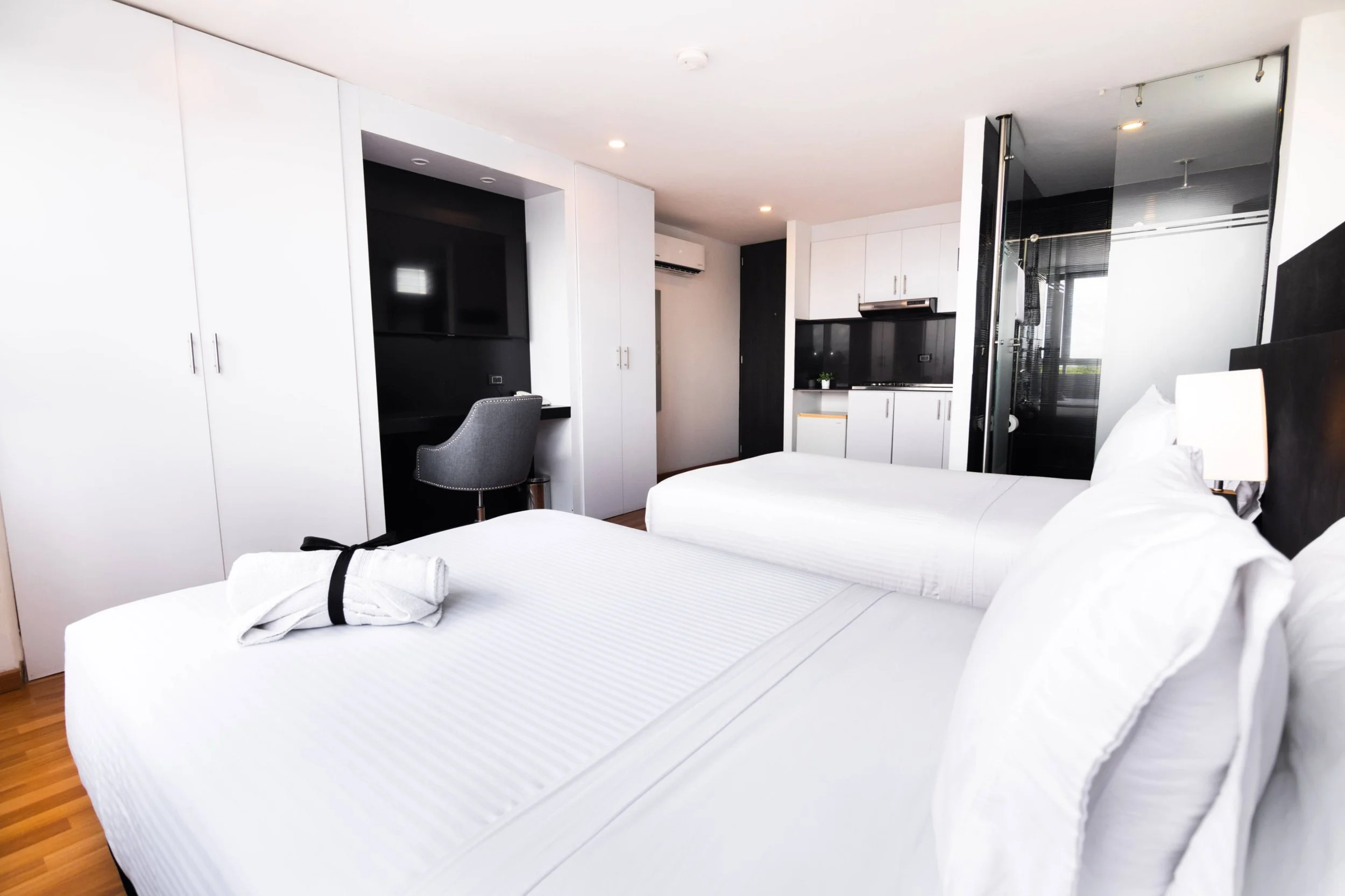 Habitación con dos camas blancas, escritorio con silla y televisión, kitchenette y baño con cabina de ducha.