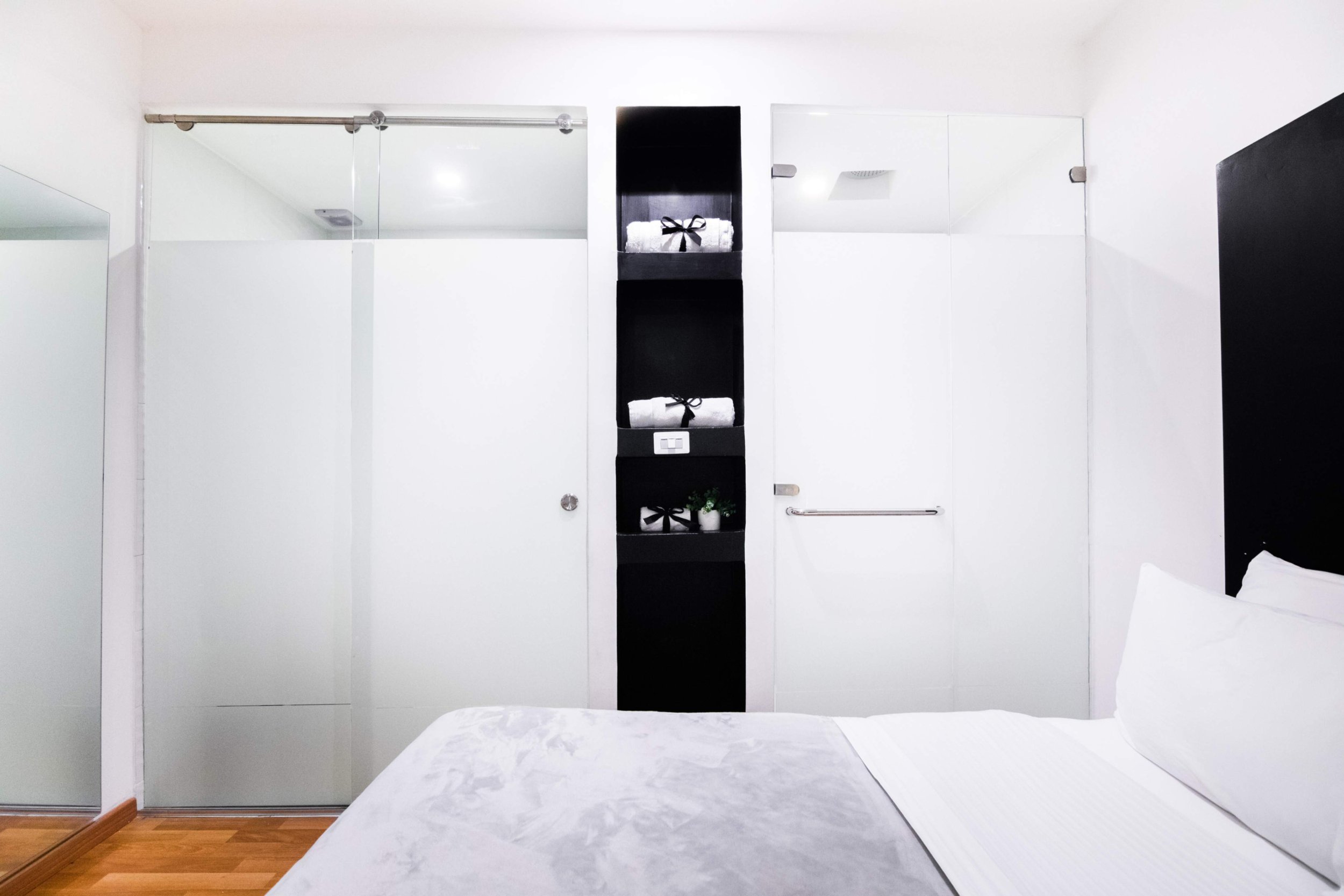 Habitación con cama, armario con puertas blancas y estantería negra con toallas blancas y planta decorativa.