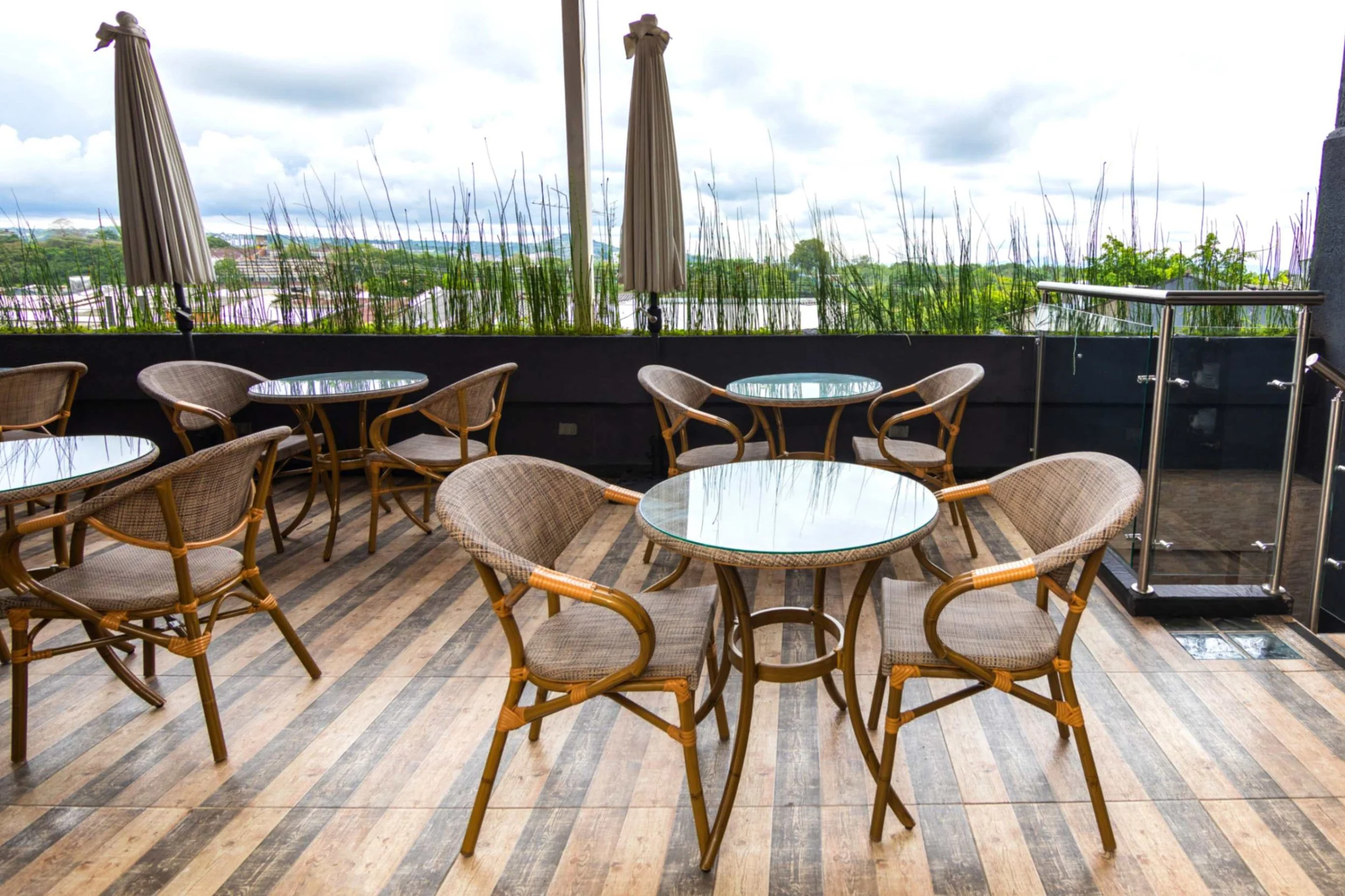 Terraza de restaurante con mesas y sillas de mimbre, sombrillas cerradas, piso de madera y vegetación alrededor.