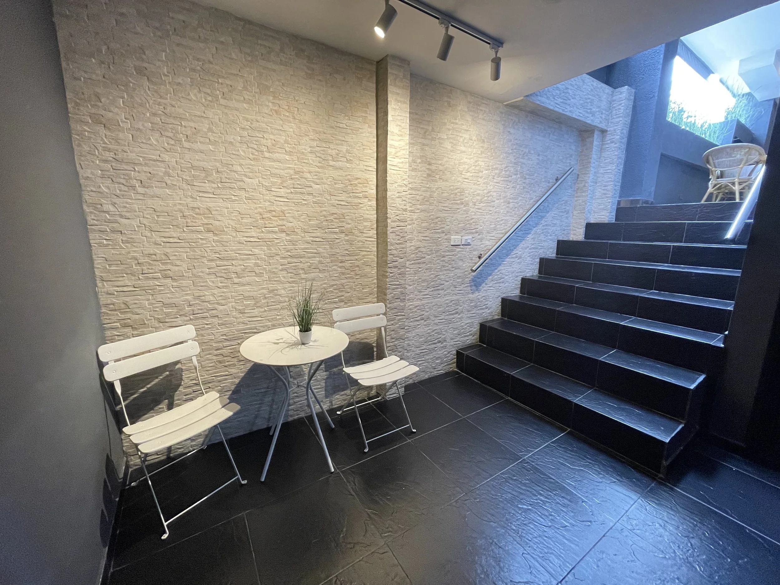 Área interior con dos sillas blancas, una mesa redonda pequeña con una planta encima y una escalera de azulejos negros a la derecha.