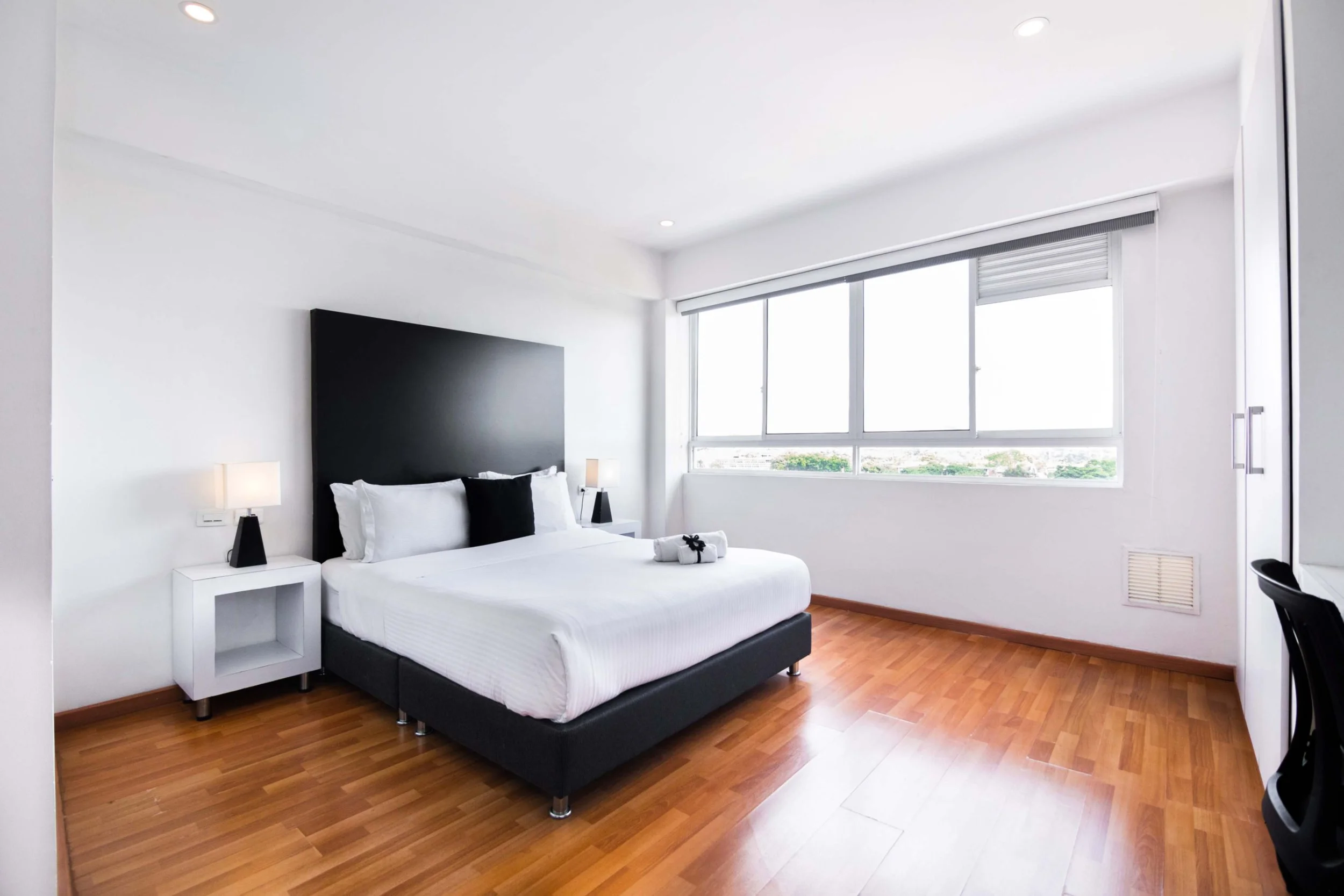 Habitación moderna con cama grande, cabecera negra, mesitas blancas con lámparas, ventanas grandes, paredes blancas y suelo de madera.