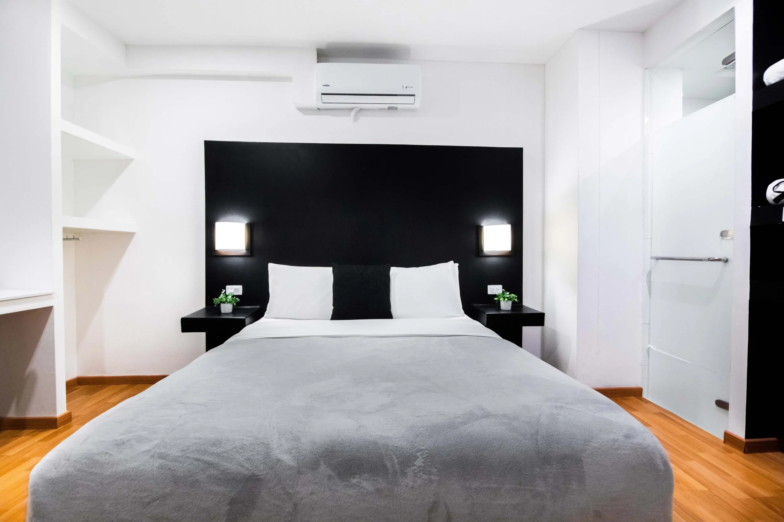 Habitación moderna con cama grande, cabecera negra, dos luces de lectura, aire acondicionado, paredes blancas, estantes blancos, piso de madera, y puerta de vidrio.