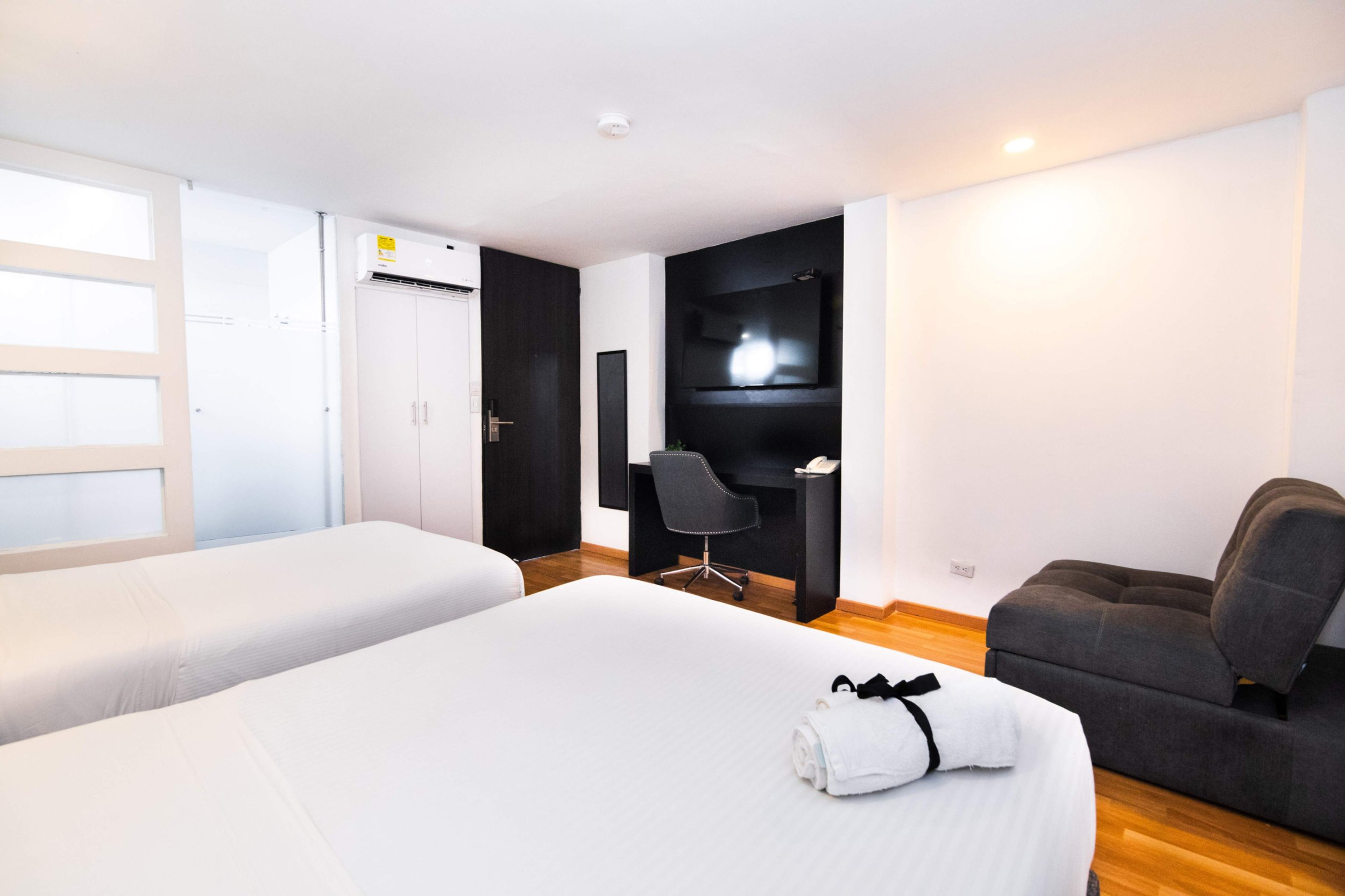 Habitación de hotel con dos camas, una toalla enrollada, silla de oficina, televisor, sillón y armario en un espacio moderno y bien iluminado.
