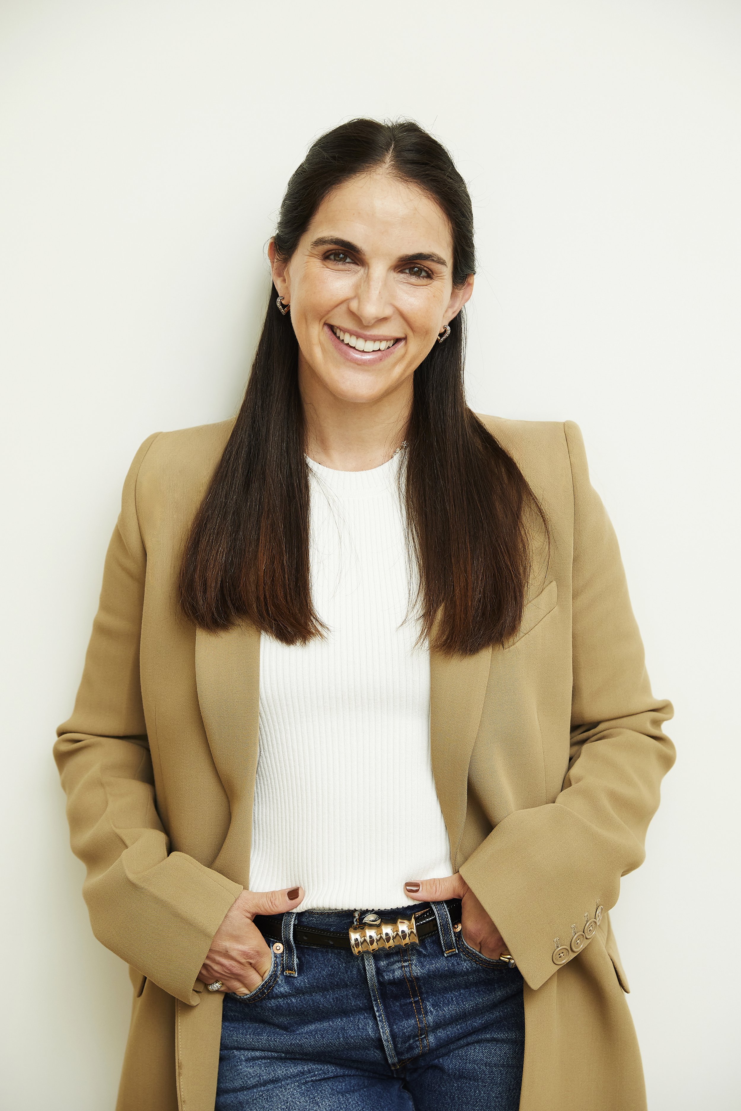 Smiling woman with long brown hair wearing a beige blazer, white top, and blue jeans, standing against a plain white wall.