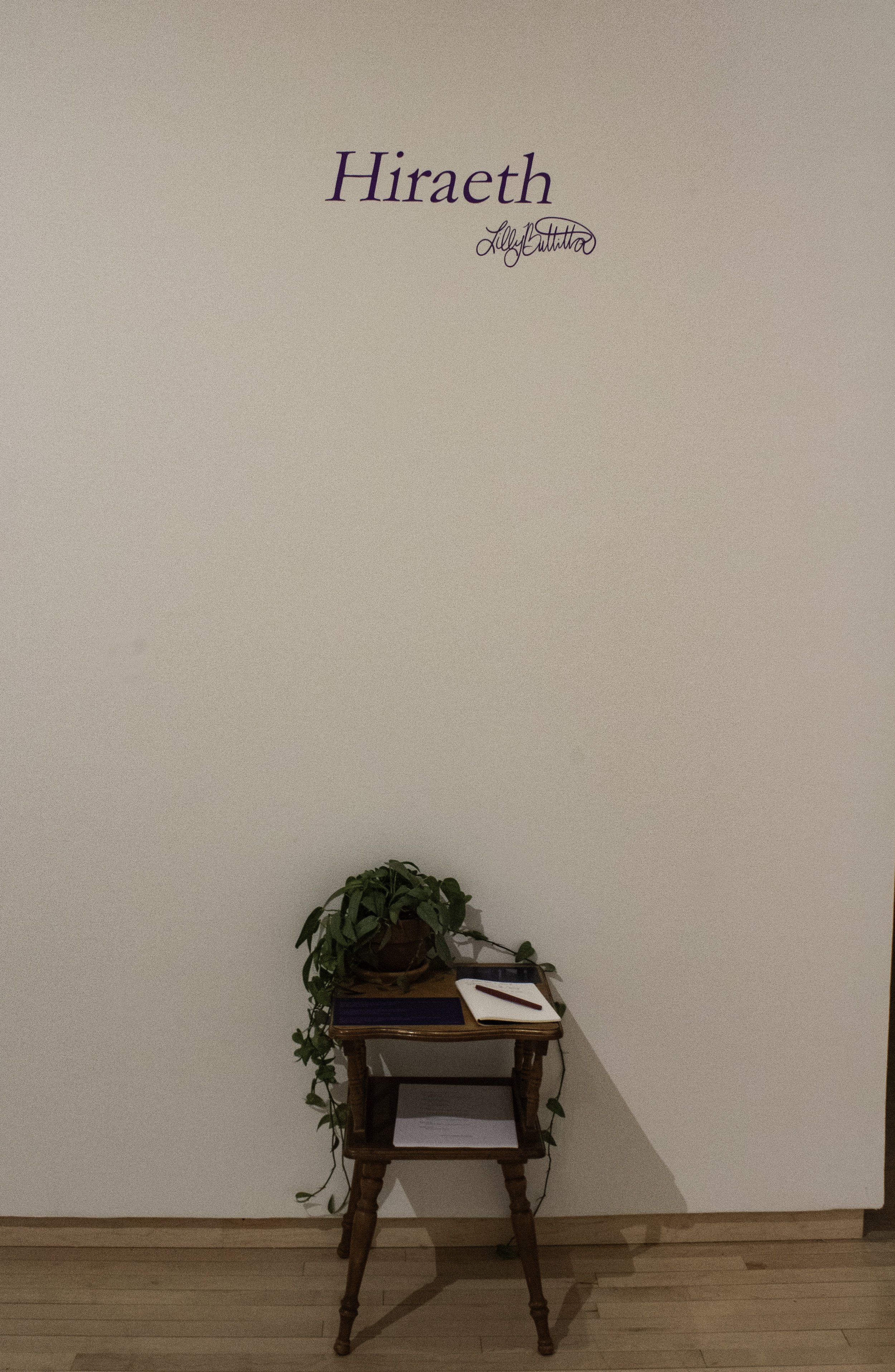 "Hiraeth," Installation view, guest book, and thesis Statement card on table with Wall Title, approx. 1 ½' x 2', 2025, Photo Credit: Lilly Buttitta