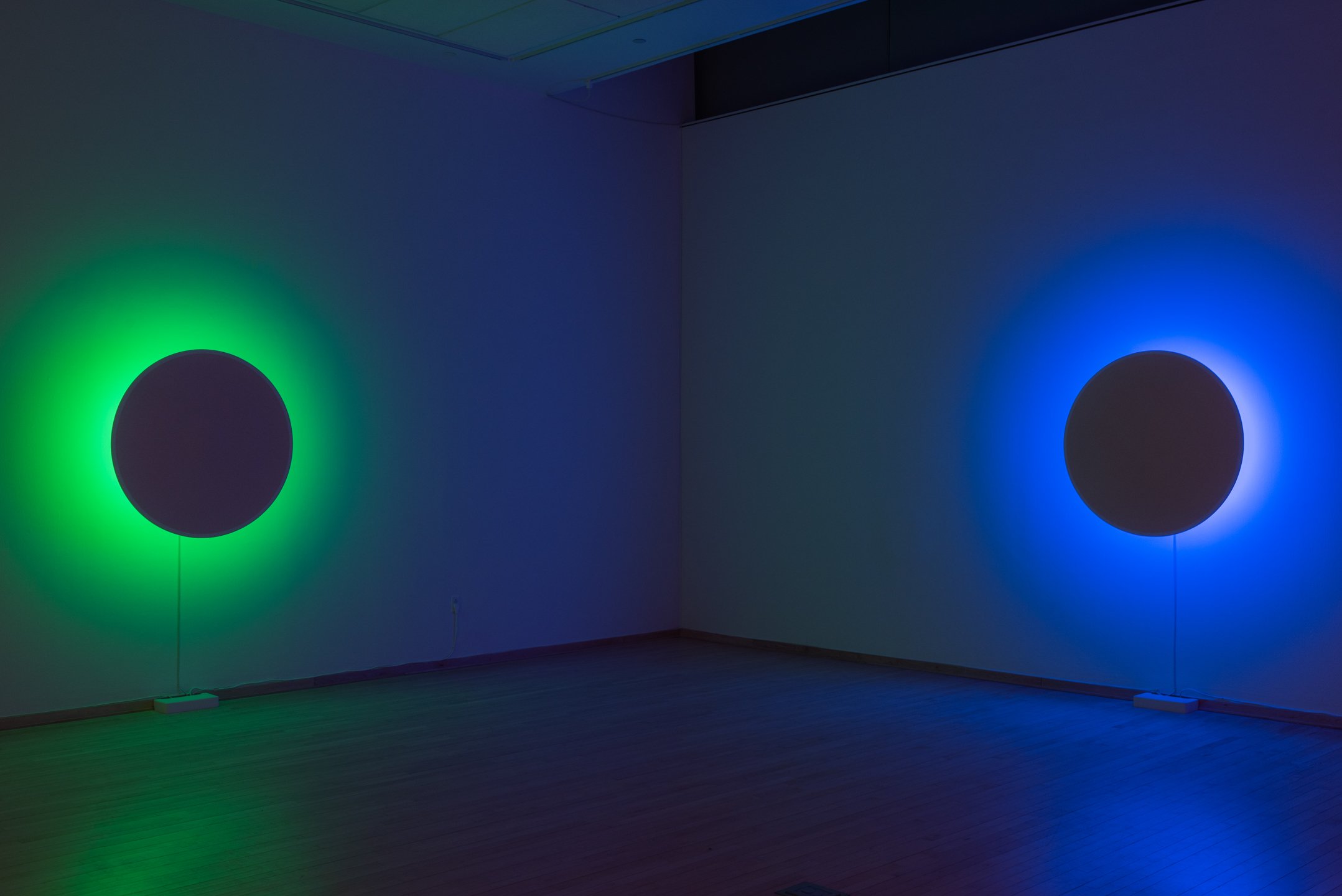"In Totality" (still), 2025, Time-based installation view showing “Optic Disc (Green)” and “Optic Disc (Blue),” Retroreflective glass microspheres, acrylic paint, foam, steel, LED light, computer controller. 46” by 46” by 14” each, Photo credit: Neig