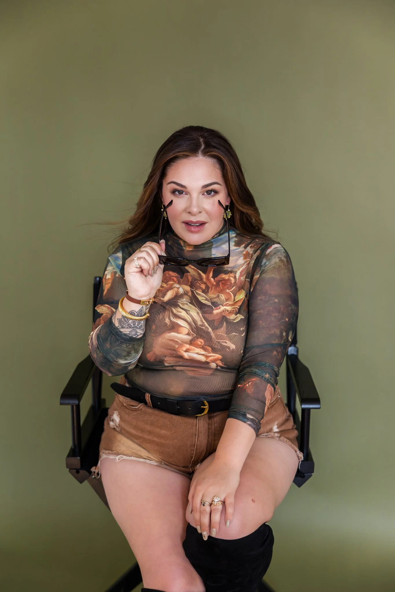 A woman with long brown hair sitting in a director's chair, holding sunglasses near her face, wearing a sheer top with a classical painting print, brown shorts, and black knee-high boots, against a plain green background.
