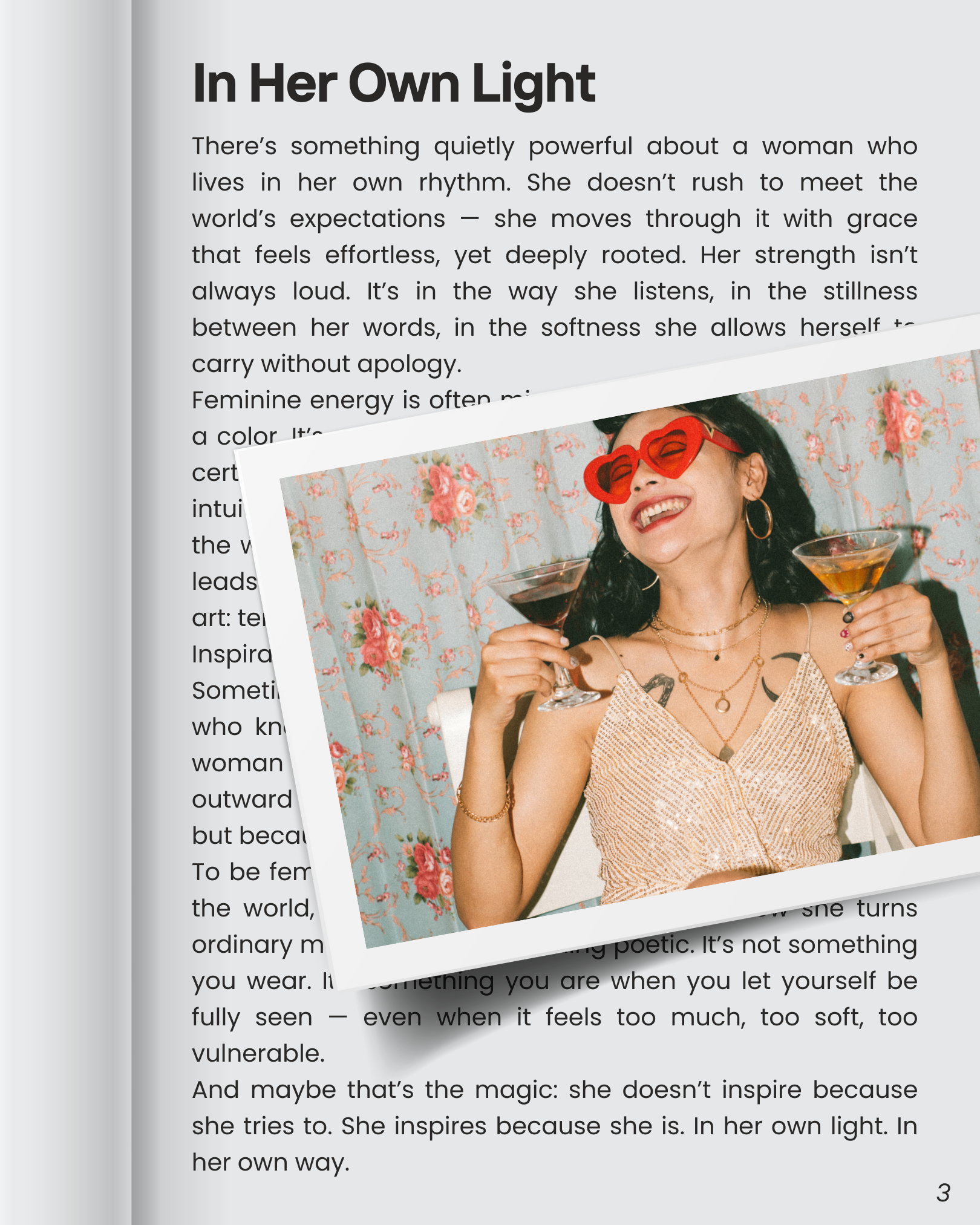 A woman wearing heart-shaped red sunglasses, gold jewelry, and a sparkly top, smiling and holding two martini glasses filled with dark red and amber drinks, standing in front of patterned floral wallpaper.