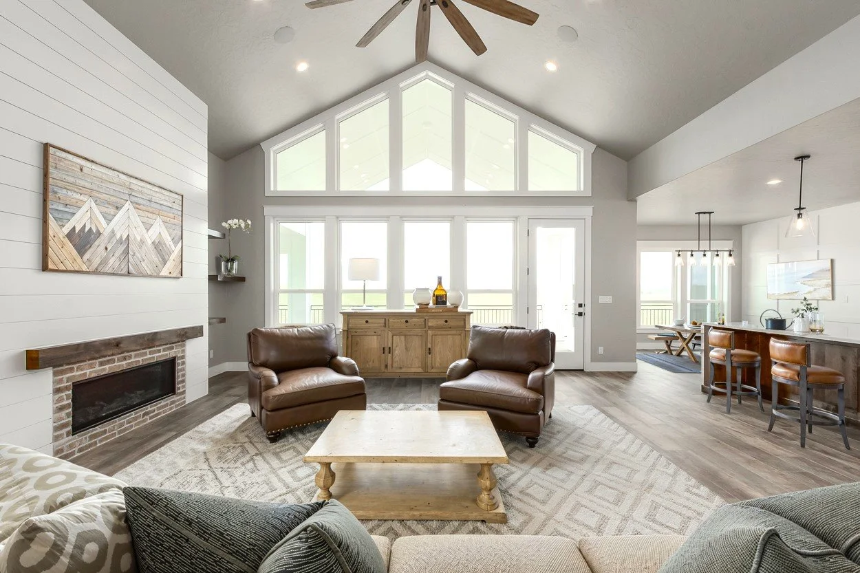 A spacious living room with a white shiplap fireplace, wooden accents, and large windows letting in natural light. There are two brown leather armchairs, a light wood coffee table, and cozy seating with pillows. The open floor plan includes a dining area and a kitchen with a bar counter and stools, and a ceiling fan with wooden blades.