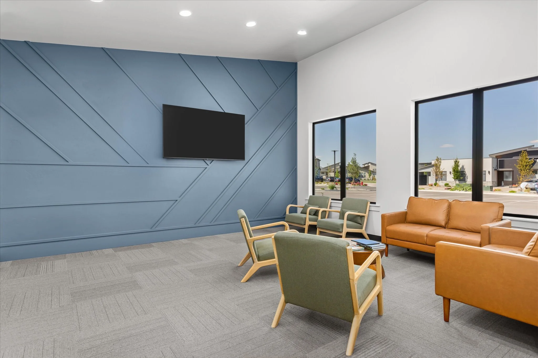 Modern waiting room with green and tan chairs, a tan sofa, large windows, blue accent wall with geometric pattern, and a flat-screen TV, in a bright and clean space.