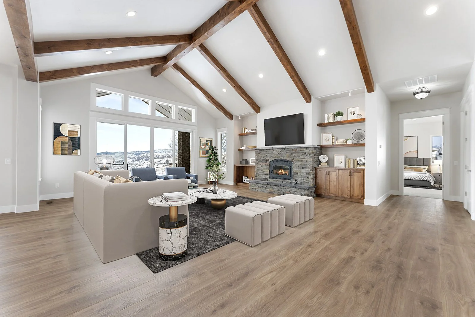 Spacious living room with high white ceiling, wooden beams, large window with mountain view, beige sofa, modern coffee table, stone fireplace with mounted TV, built-in shelves with decor, and view into an adjacent bedroom.