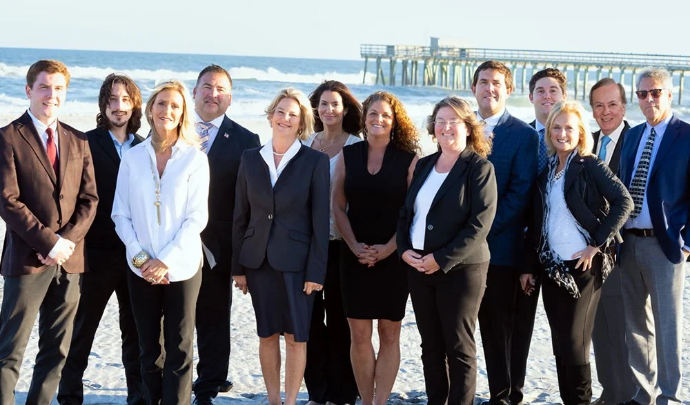 Group of professionally dressed people posing on a beach with ocean and pier in background.