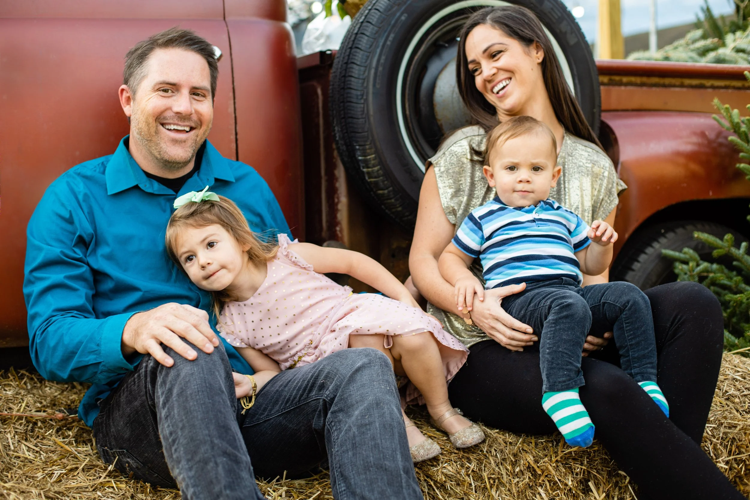 A happy family of four sitting on hay outdoors with a rusty truck in the background. The father is wearing a blue shirt, the mother is smiling with long dark hair, the young girl is leaning on the father wearing a light pink dress with a green bow, a