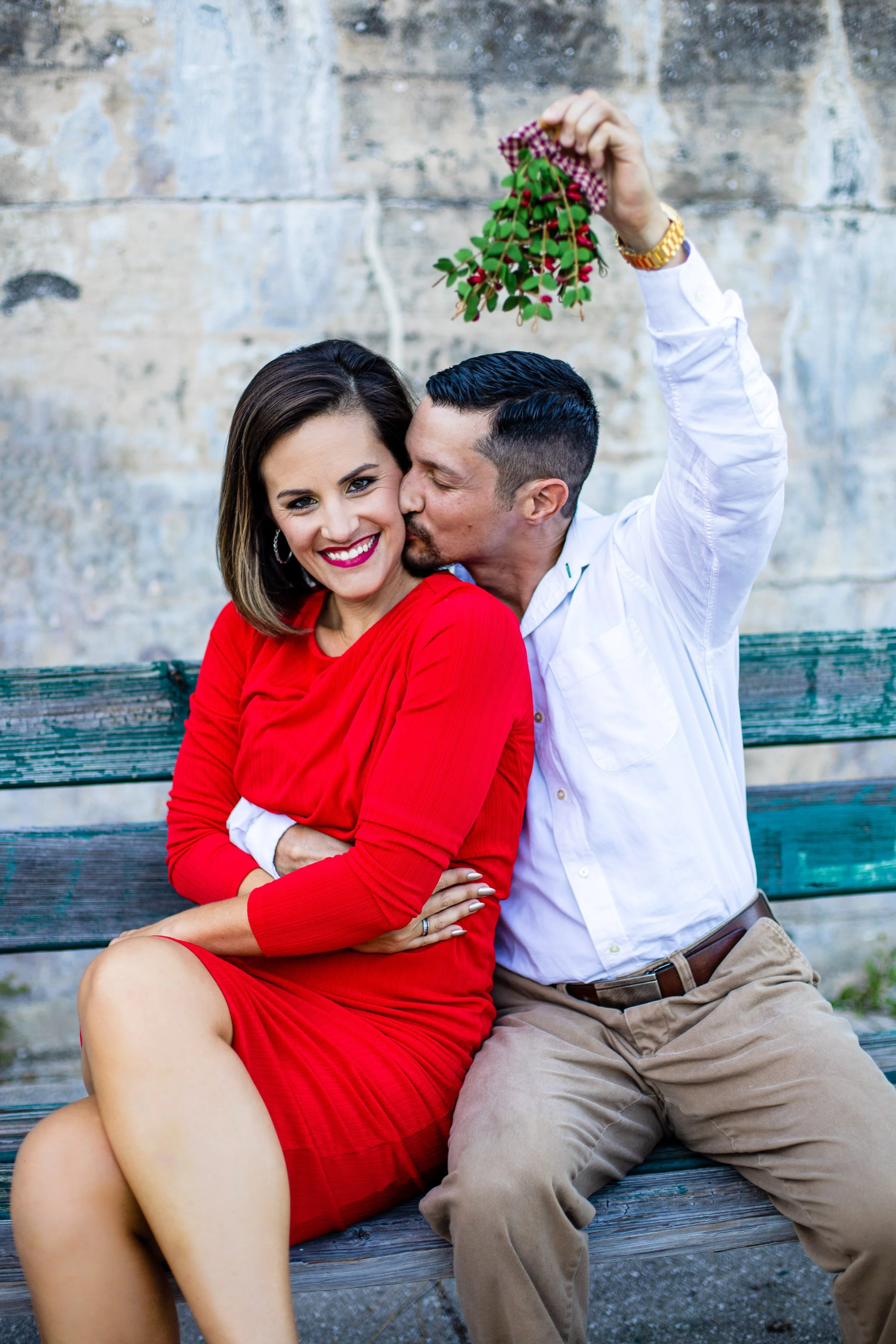 A man and woman sit on a park bench, with the man kissing the woman's cheek as she smiles. The woman wears a red dress, and the man is dressed in a white shirt and khaki pants, holding a small branch with red berries and green leaves above their head