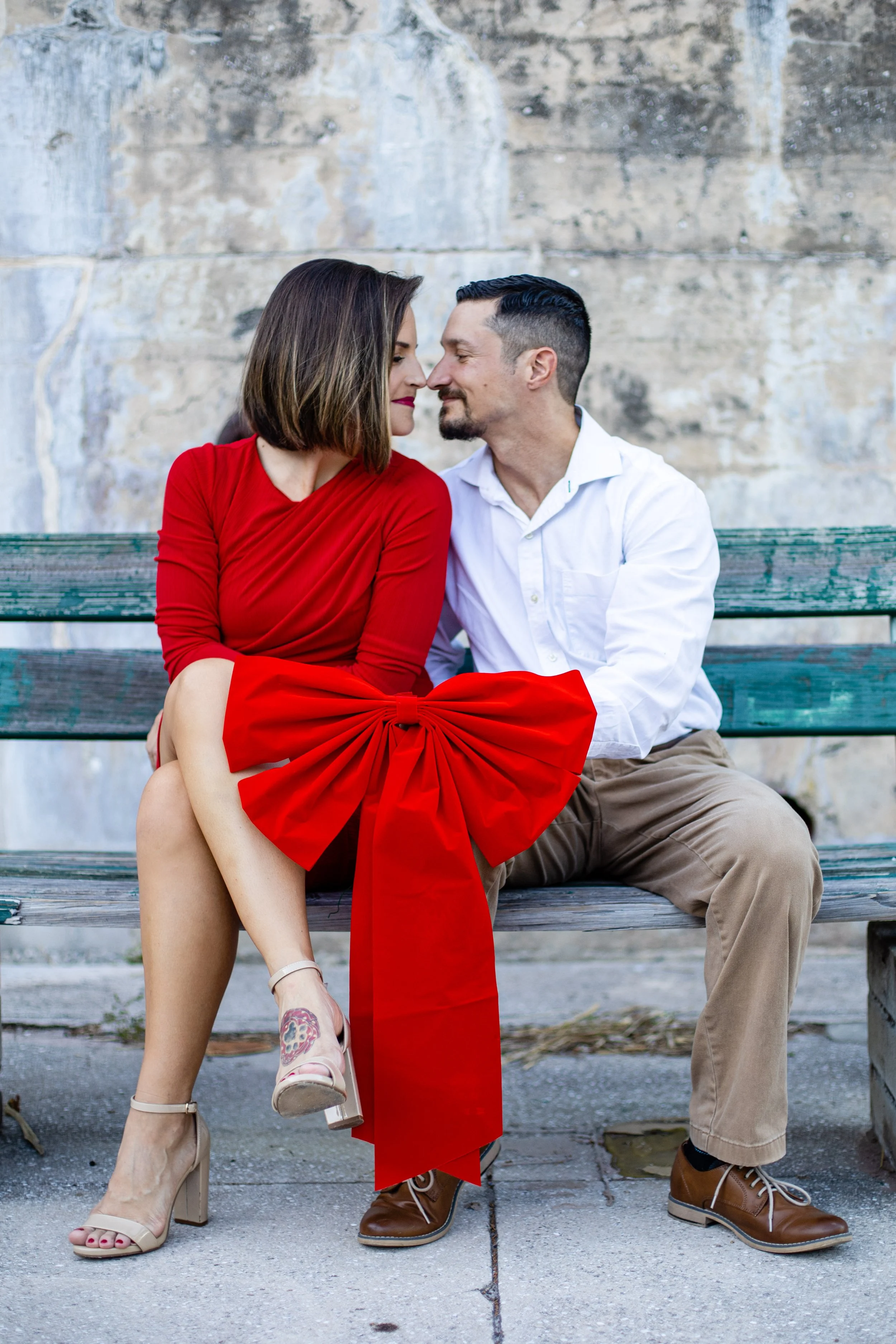 A man and woman sitting closely on a park bench, touching foreheads, with a large red bow on the woman's lap, both dressed semi-formally.