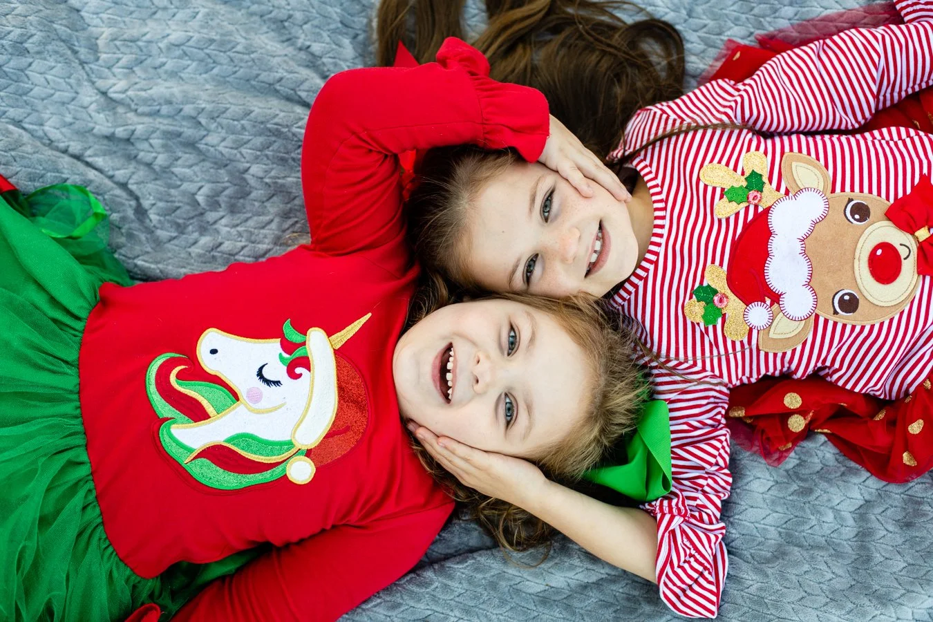 Two young girls lying on a gray quilted blanket, wearing Christmas-themed pajamas with unicorn and reindeer designs, smiling and looking at the camera.