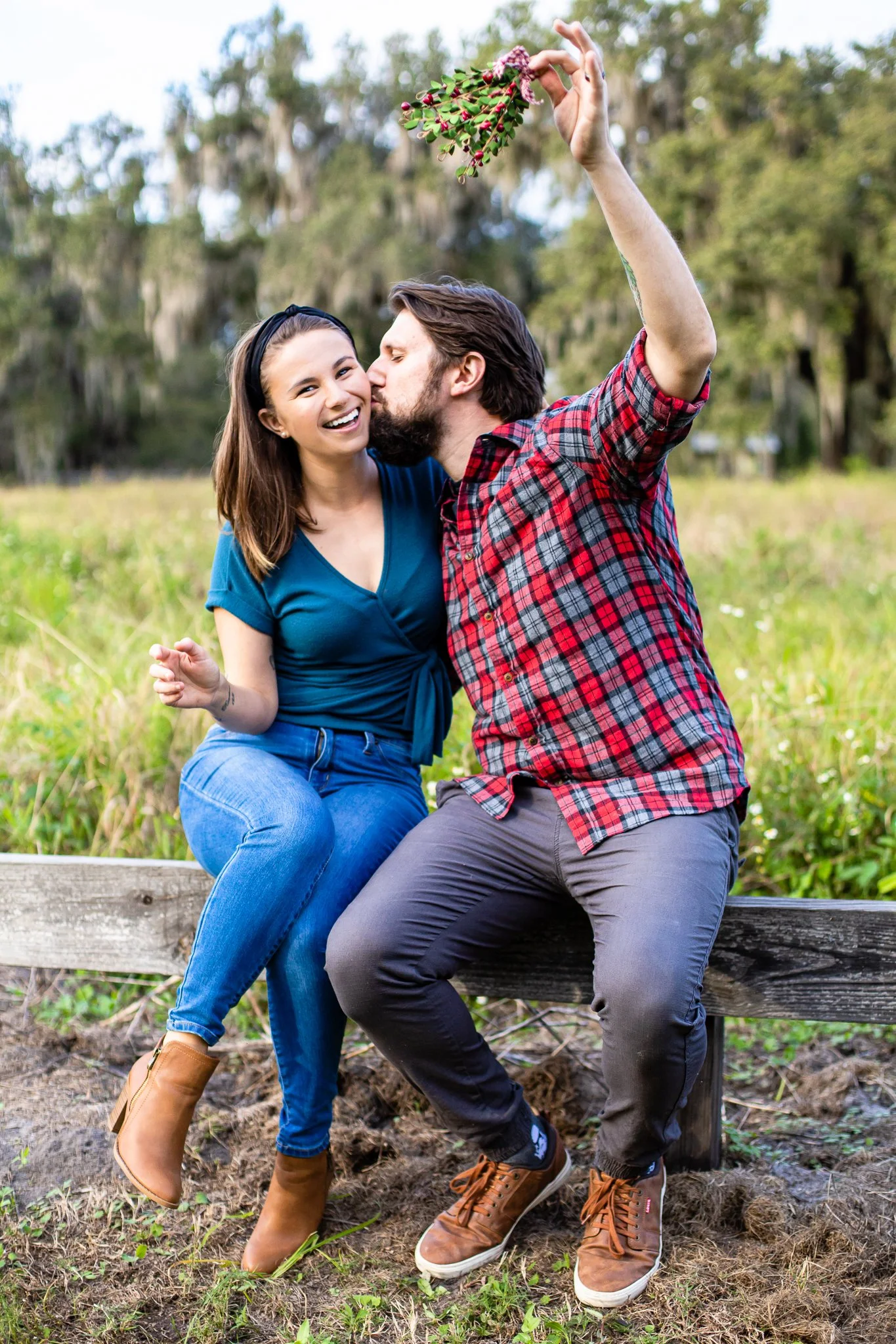 A couple sitting on a wooden bench outdoors, with the woman smiling and the man kissing her cheek while holding a bouquet of pink and green flowers.
