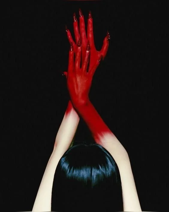 A person with black hair holds their hands together, with one hand painted red and the other white, against a black background.