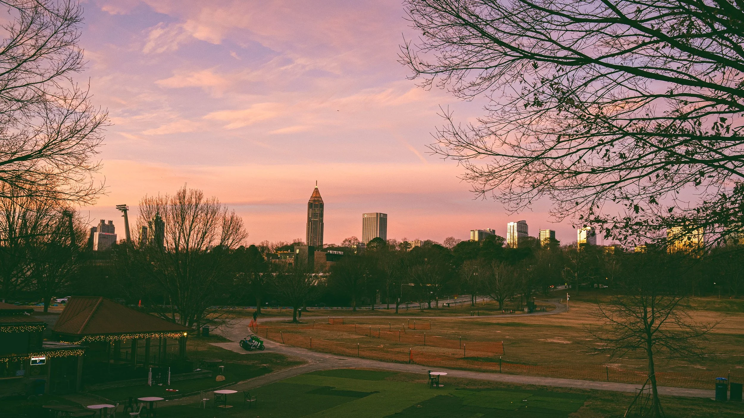 A city skyline at sunset with tall buildings, leafless trees in the foreground, and a park area with walking paths and a gazebo.