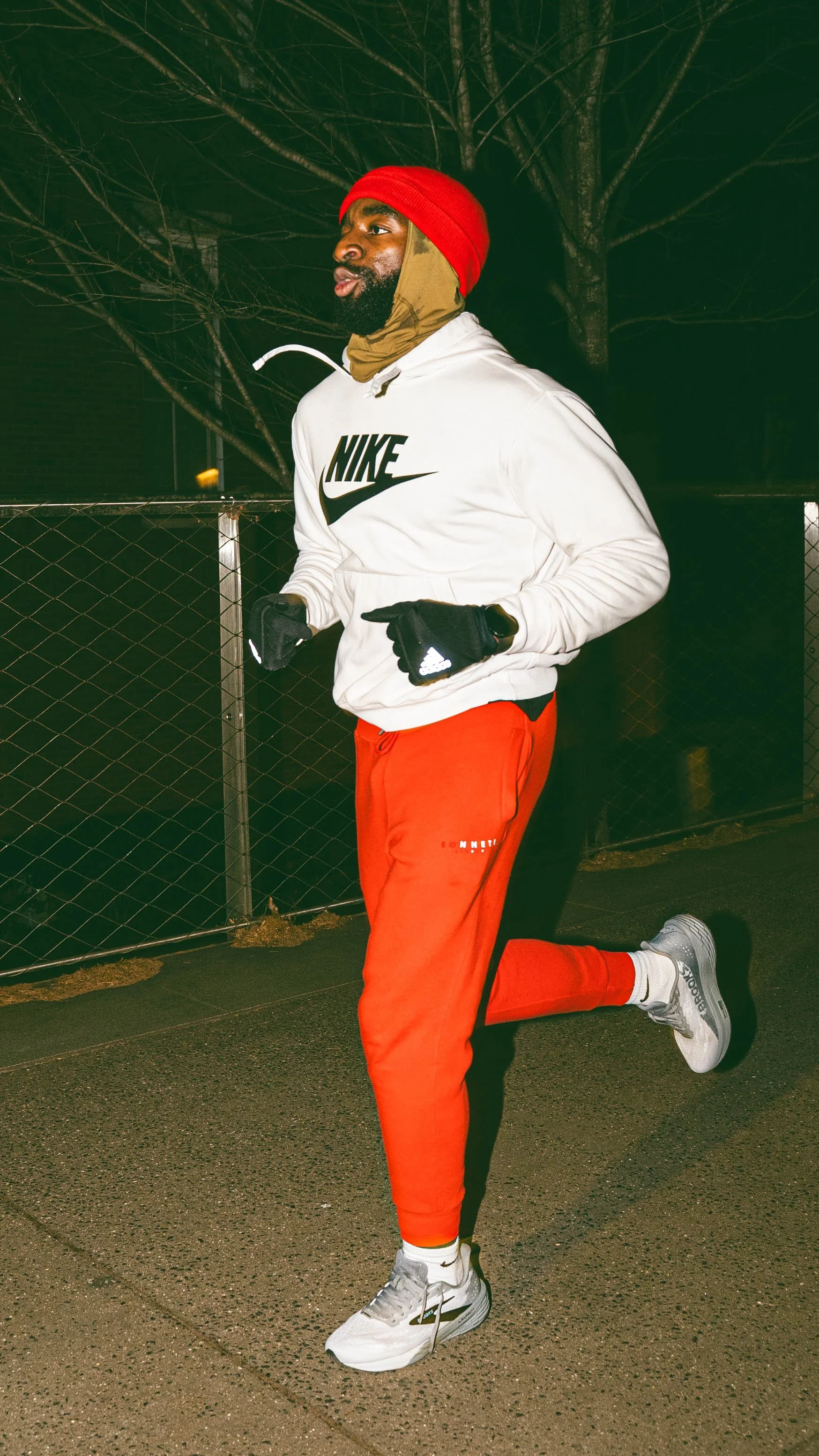 A man jogging outdoors at night, wearing a white Nike hoodie, red pants, gray and white running shoes, a red beanie, and gloves.