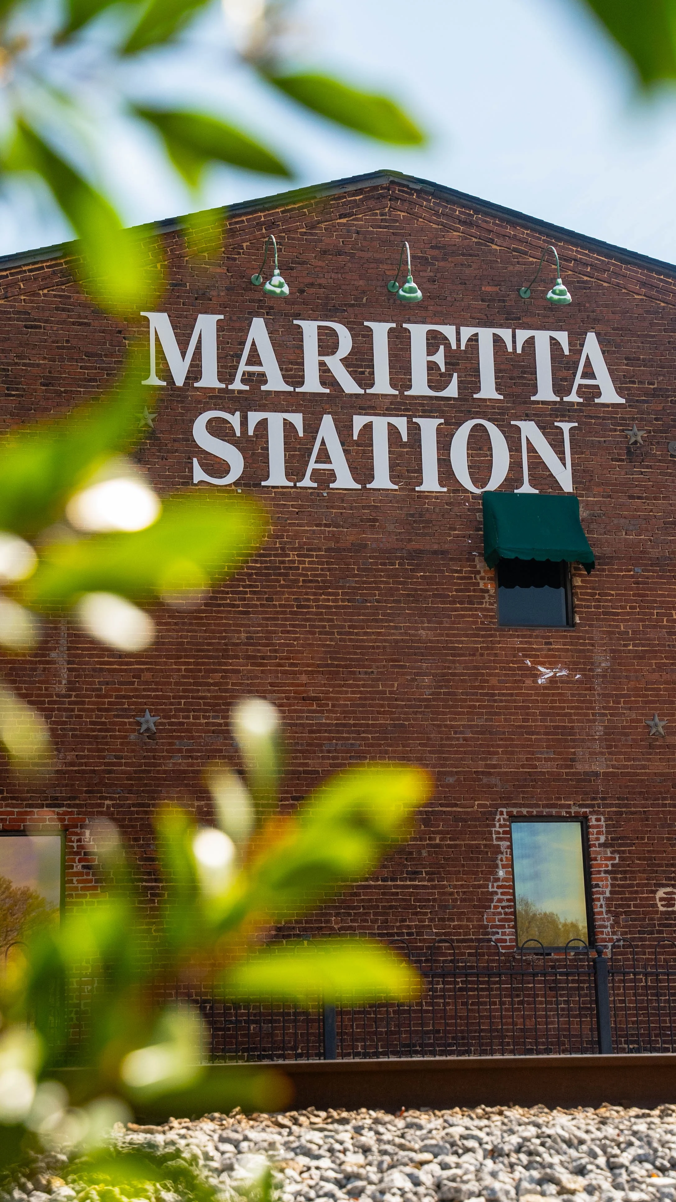 Brick building with large white sign reading 'Marietta Station' and green outdoor lights, partially obscured by leaves in the foreground.