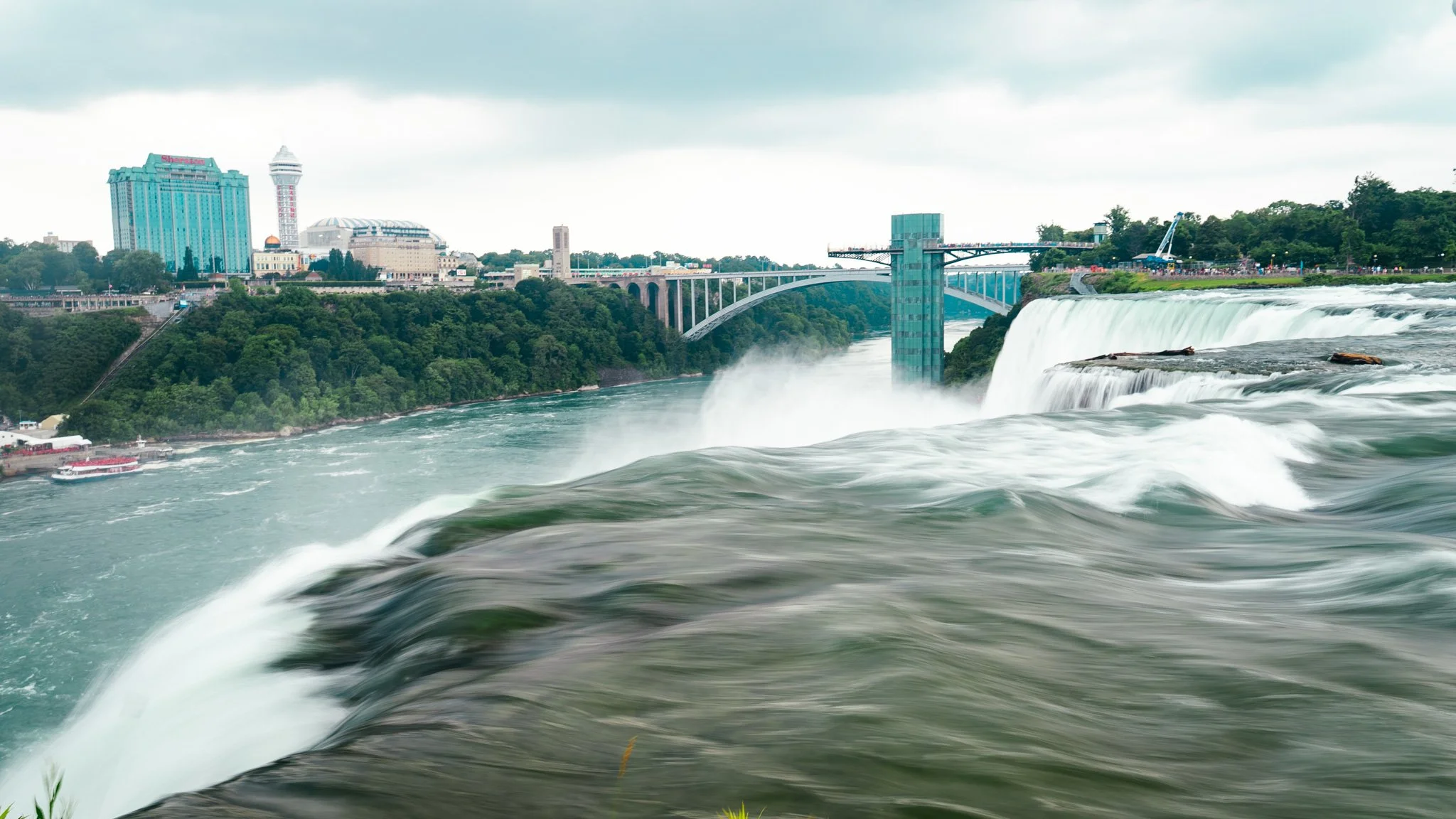 Niagara Falls with the city skyline in the background, including a tall blue building and a viewing tower, under cloudy skies.