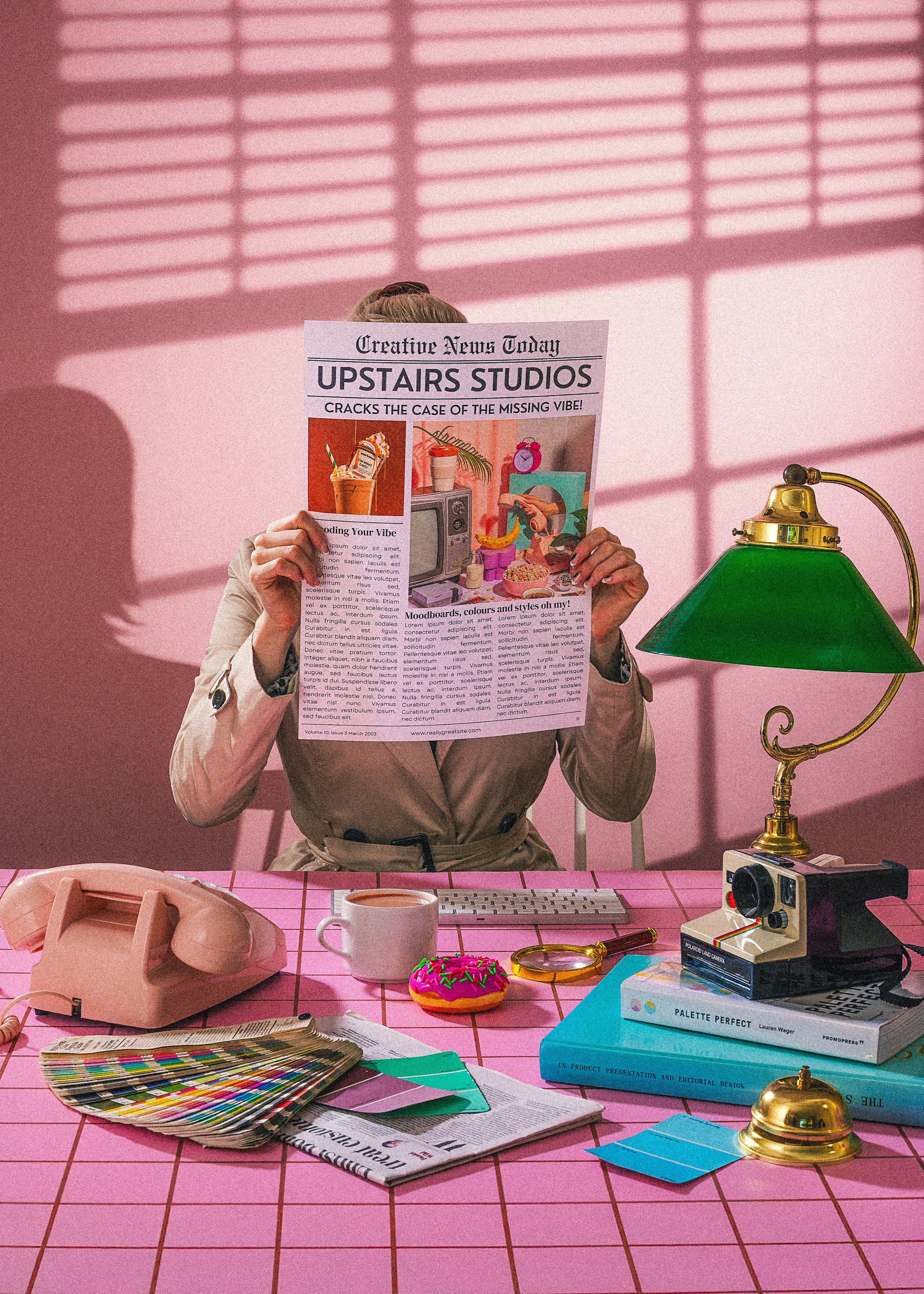 A woman sitting at a pink desk, holding a newspaper in front of her face, in a room with pink walls. The desk has a pink rotary phone, a coffee mug, a colorful fan deck, several papers, a small stack of books, a vintage camera, and a gold desk bell. A green desk lamp is on the right side of the desk. The woman is wearing a beige coat with the sleeves rolled up.