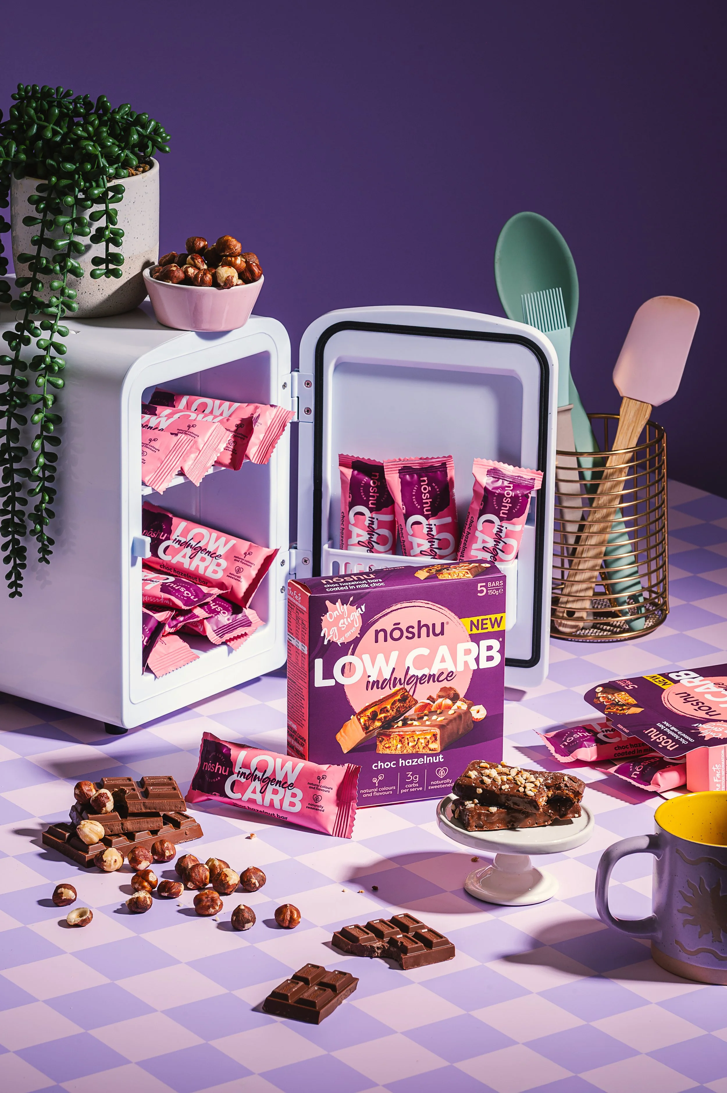 An assortment of low-carb snack bars, chocolates, and nuts displayed on a checkered table with a purple background, a potted plant, and kitchen utensils in the scene.