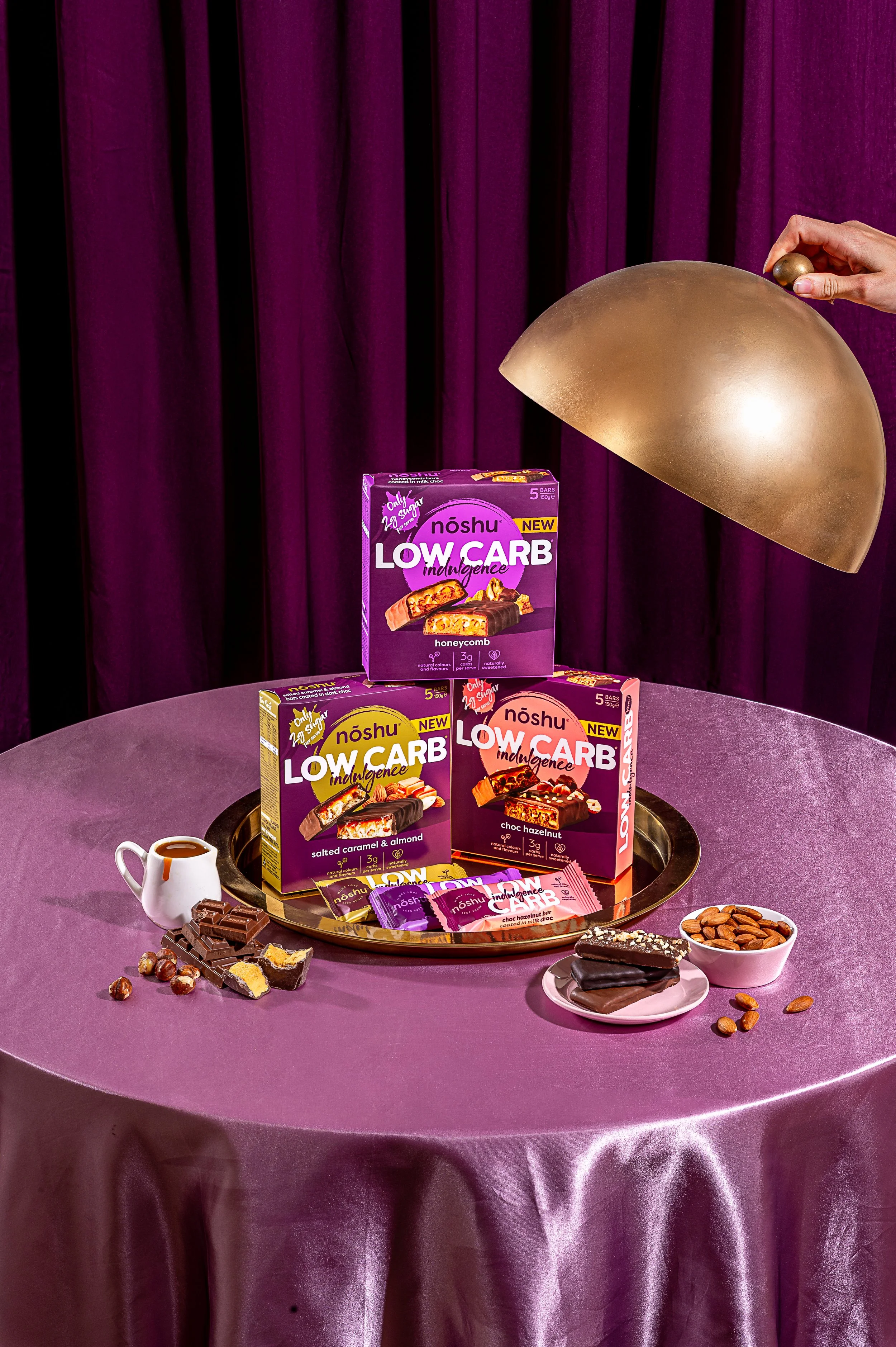 Display of Noshu Low Carb snack bars on a purple table with almonds, chocolate pieces, and a small creamer jug against a purple curtain background.