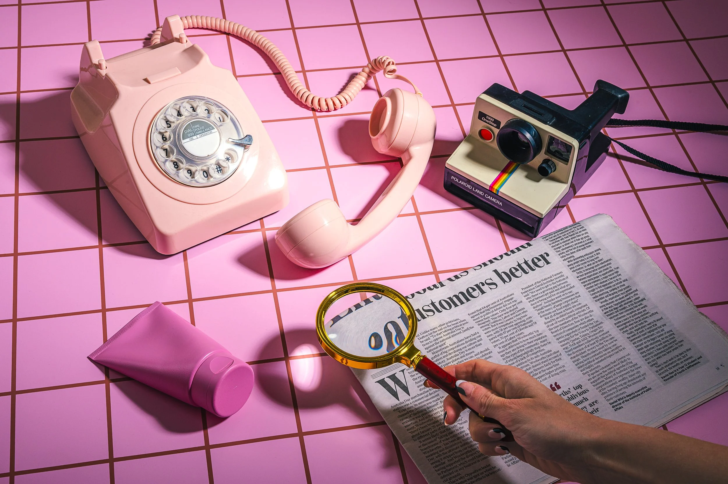A vintage pink rotary phone, a Polaroid Land Camera, a tube of pink cream, a newspaper, a magnifying glass, and a pink hairdryer are arranged on a pink tiled surface.